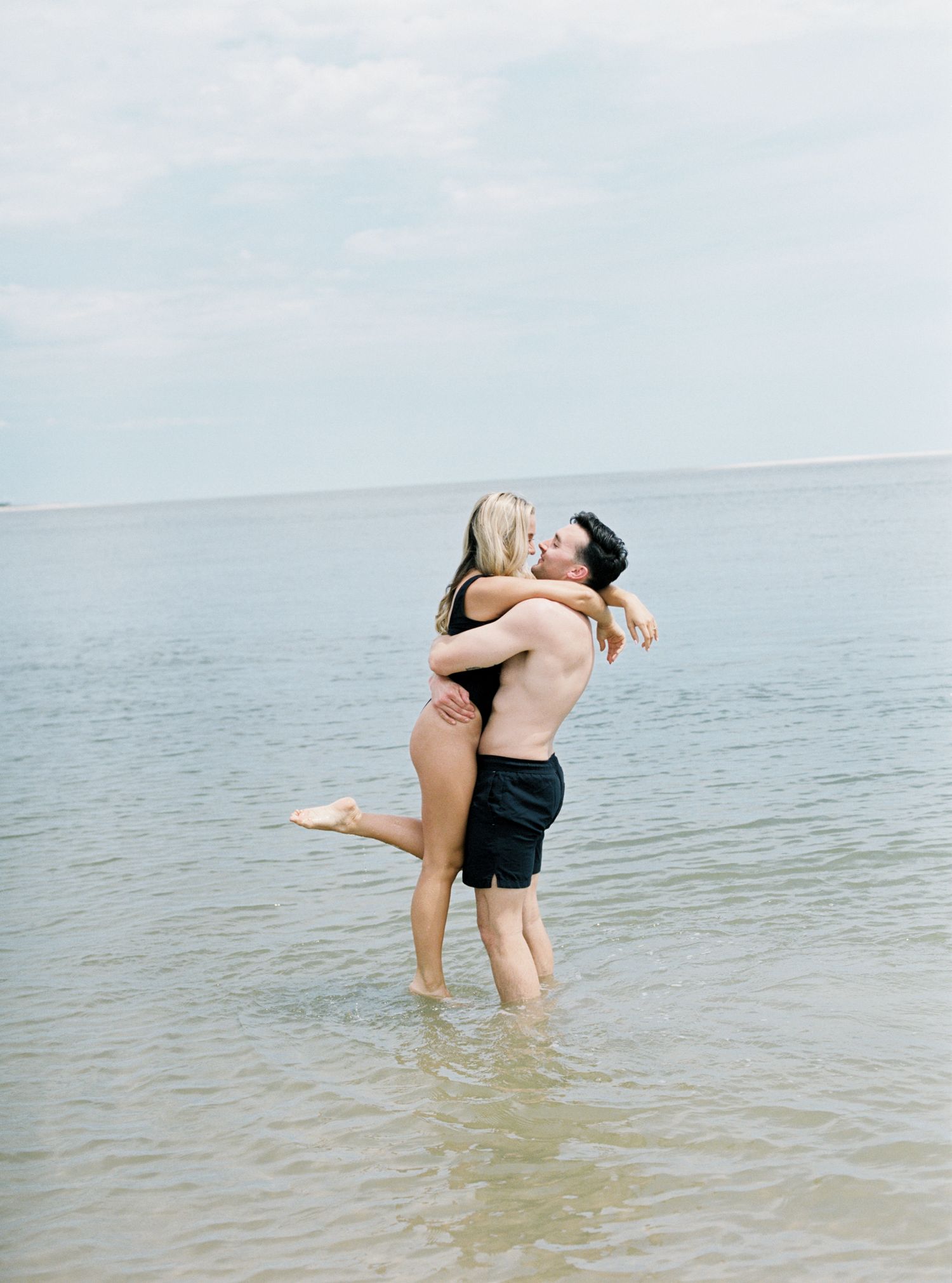 Couple embracing and laughing in shallow ocean water during romantic beach photoshoot.