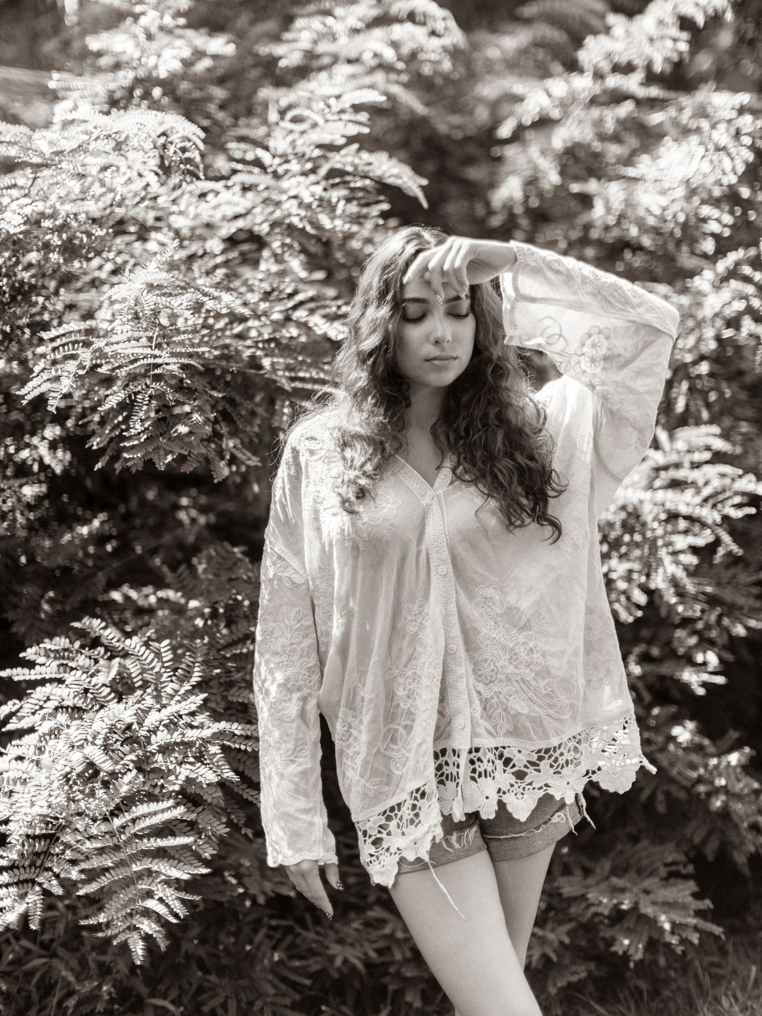 Black and white photo of a woman in lace bohemian top posing among lush ferns in a serene outdoor setting.