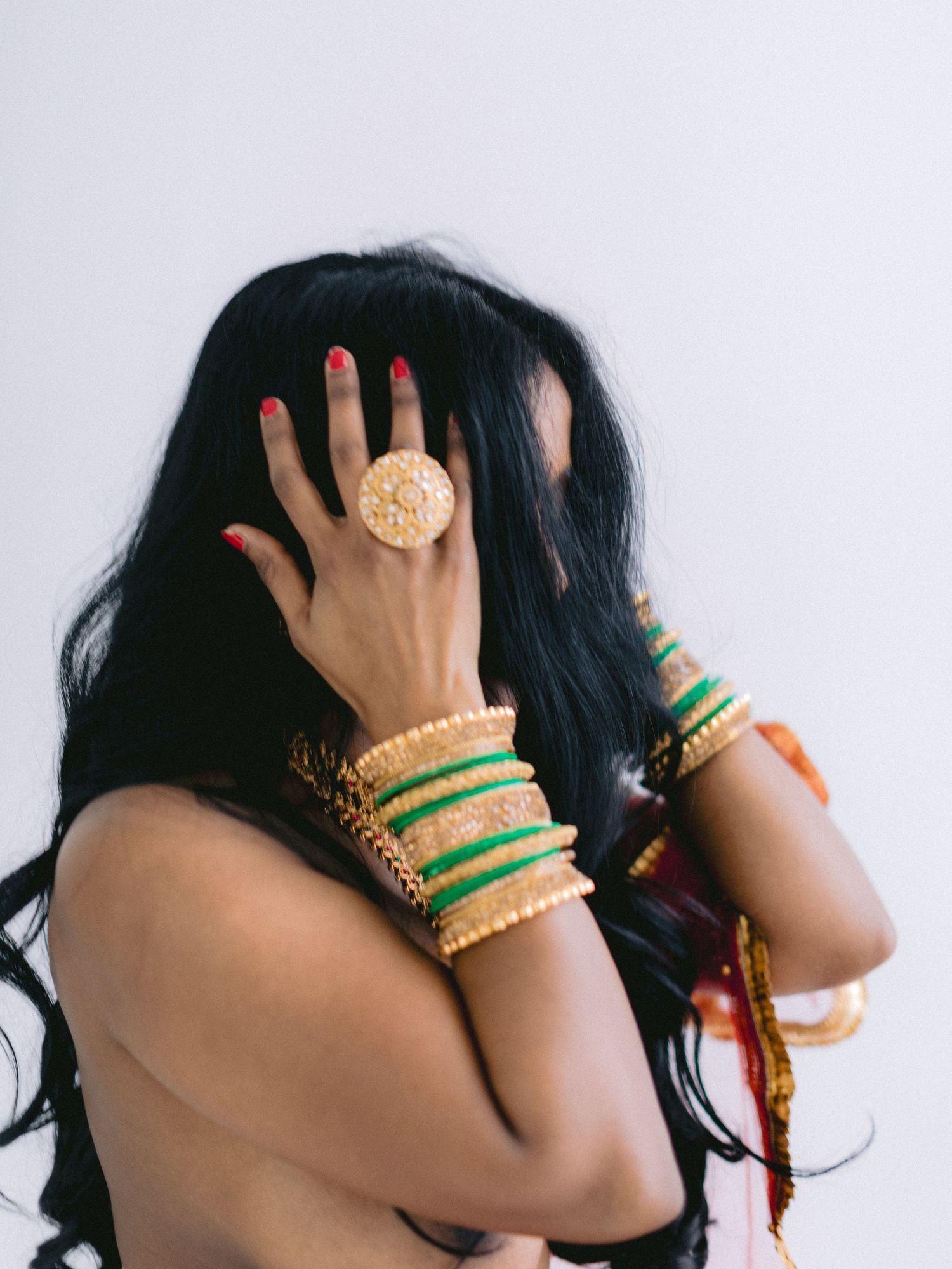 Woman adorned with gold bangles, green bracelets, and a floral ring poses with hand covering face.