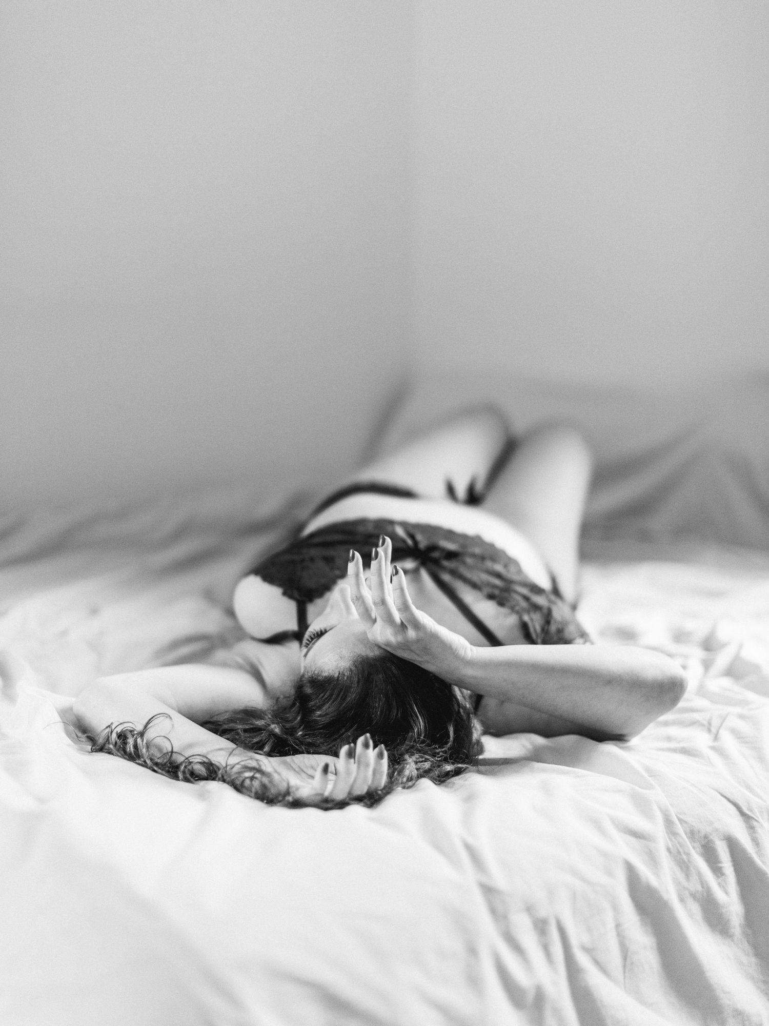 Black and white boudoir photo of a woman lying on white bedding in lingerie, covering her face with her hands.
