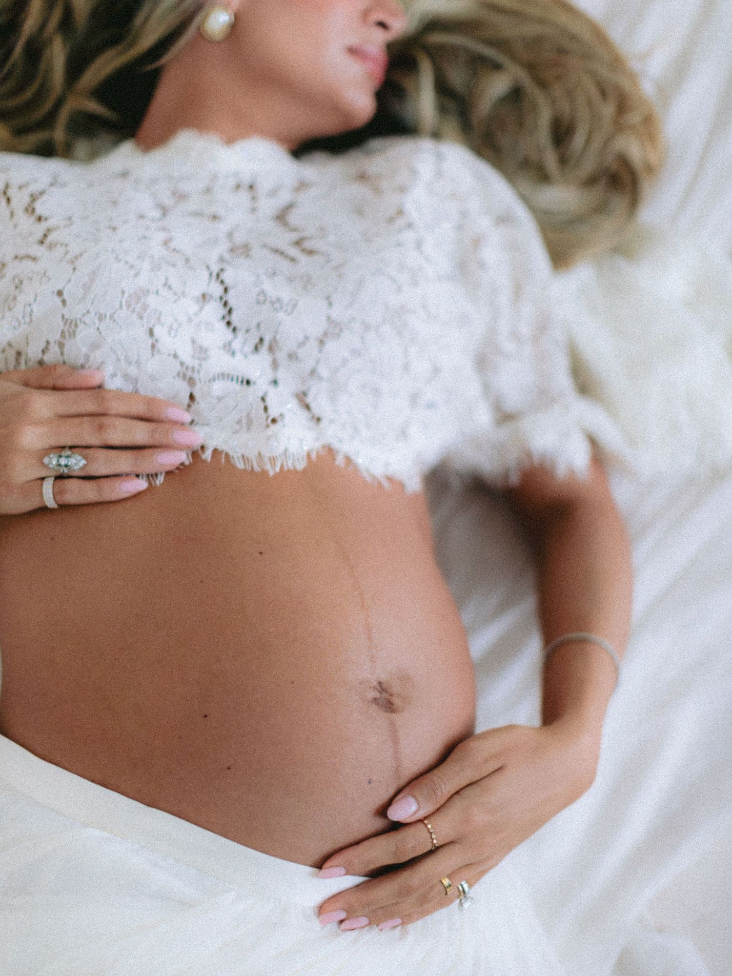 Pregnant woman in white lace crop top lying down, cradling baby bump with ringed hands.