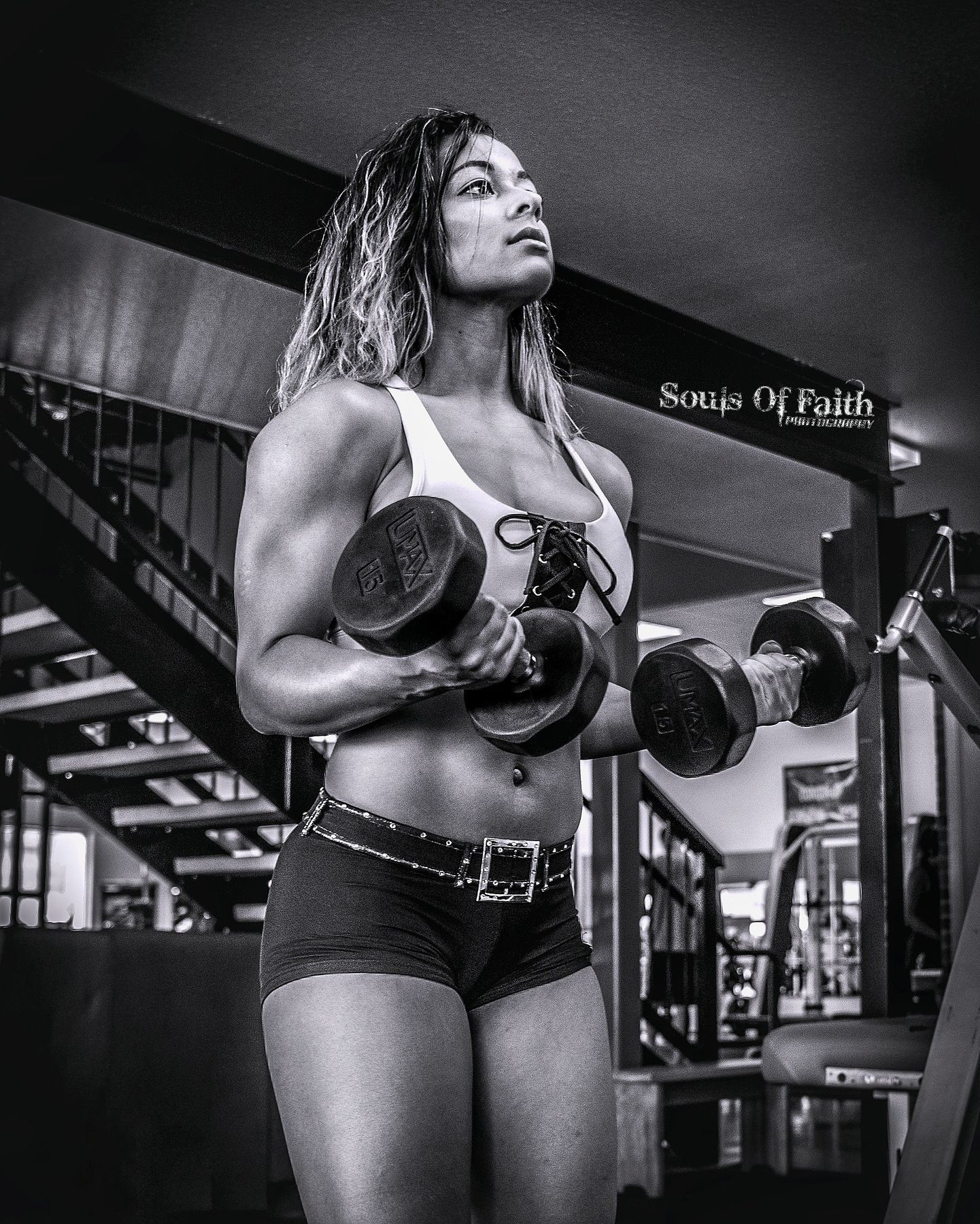 Black and white photo of a muscular woman lifting dumbbells in a gym, wearing crop top and shorts.