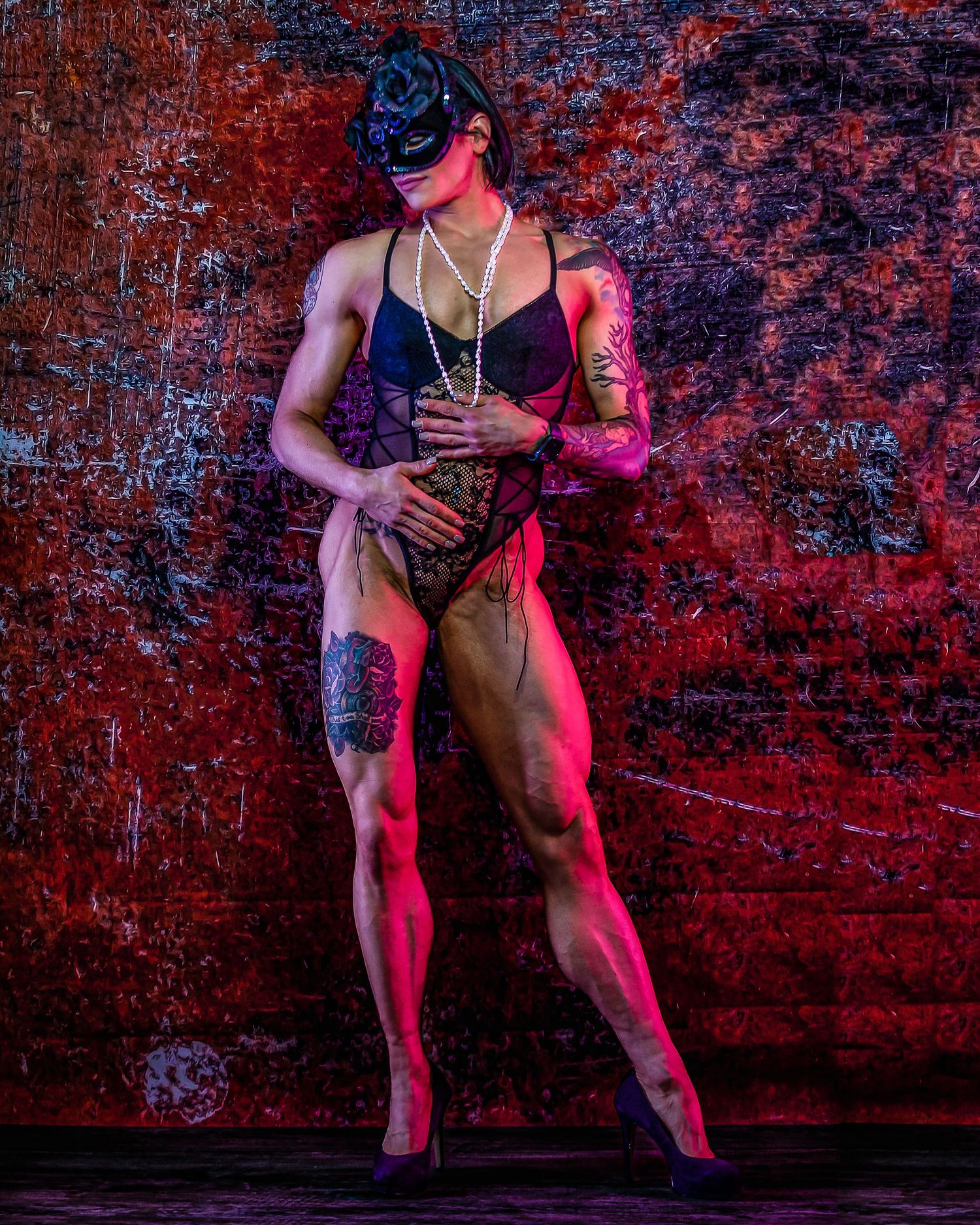 Muscular performer in black bodysuit with tattoos, pearl necklace, and masquerade mask poses under red and pink stage lighting.