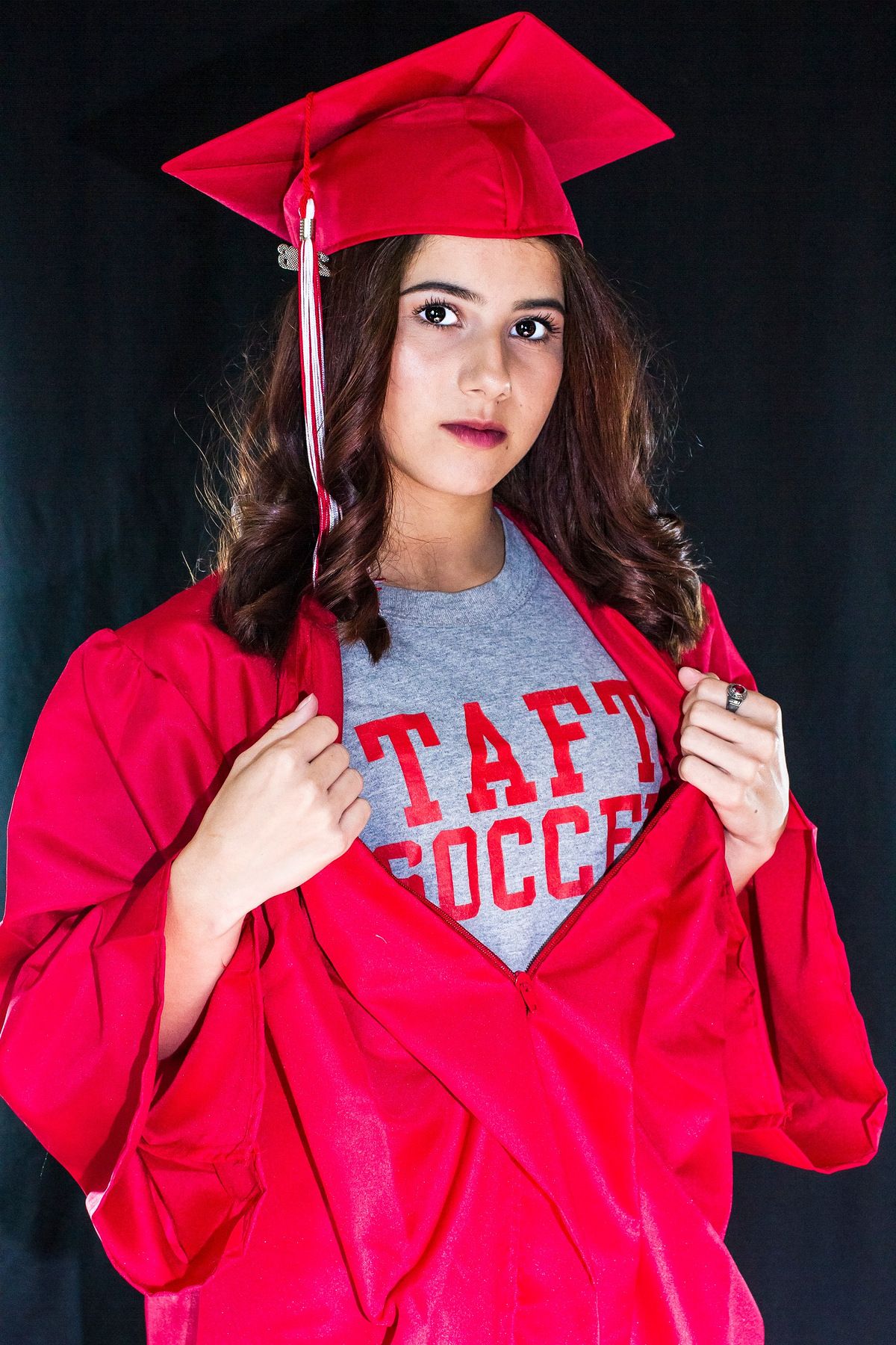 Young woman in red graduation cap and gown revealing Taft Soccer shirt, posing against dark background.