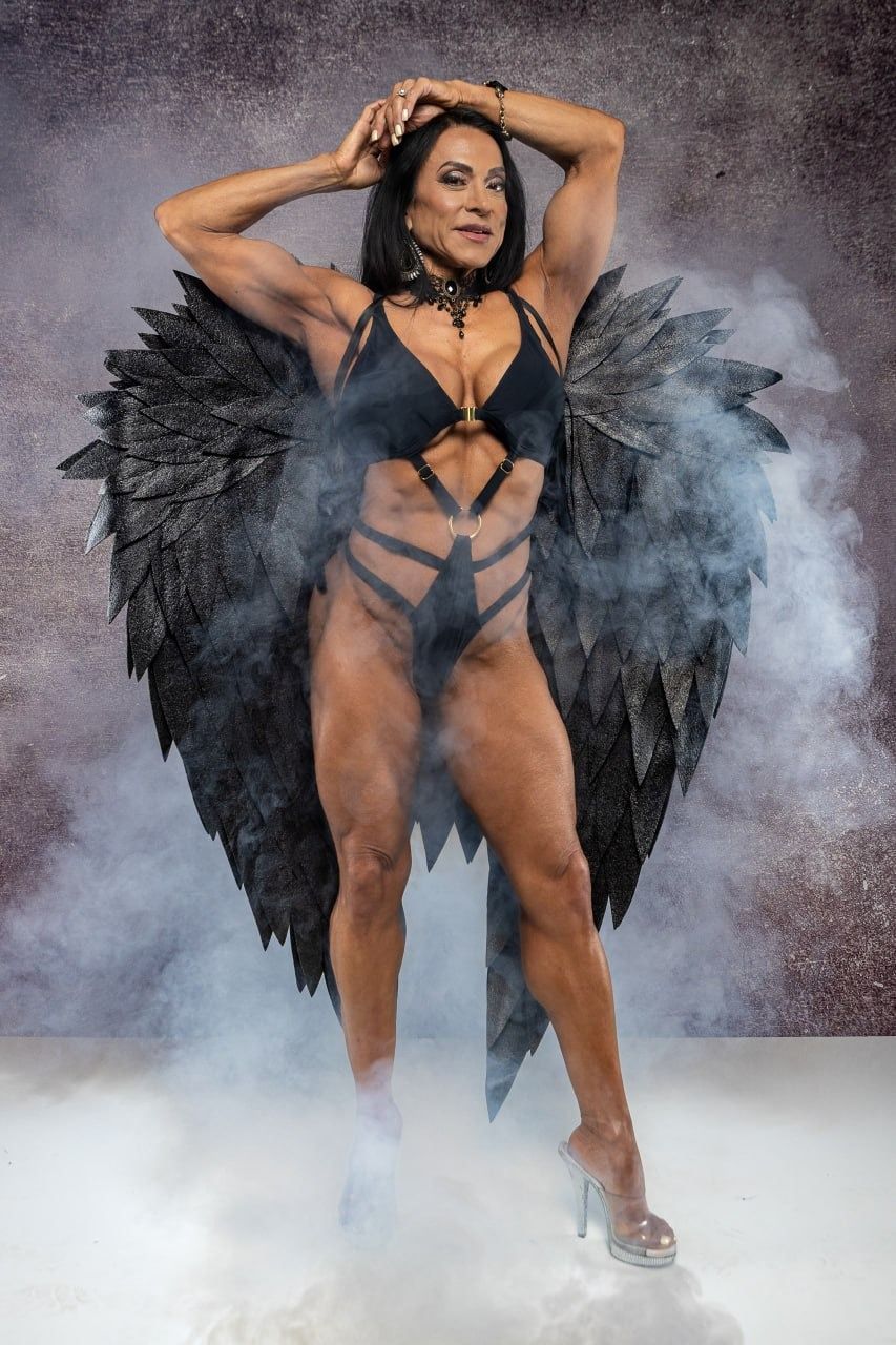 Muscular woman in black bodysuit with large dark angel wings poses dramatically in smoky studio setting wearing heels.