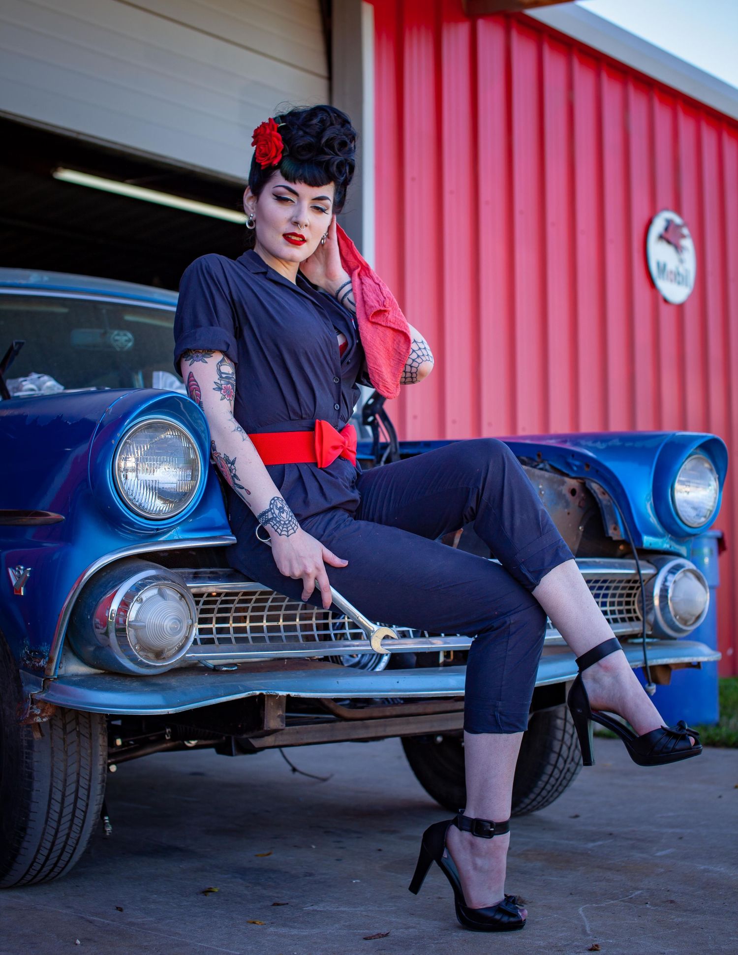 Rockabilly pin-up woman in dark jumpsuit with red belt poses on vintage blue car holding wrench outside garage.