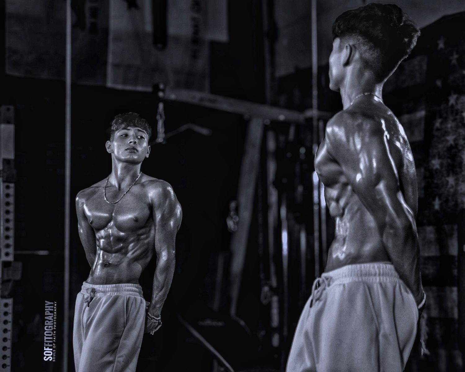 Muscular shirtless man poses in gym mirror reflection, black and white fitness photography showcasing defined physique.
