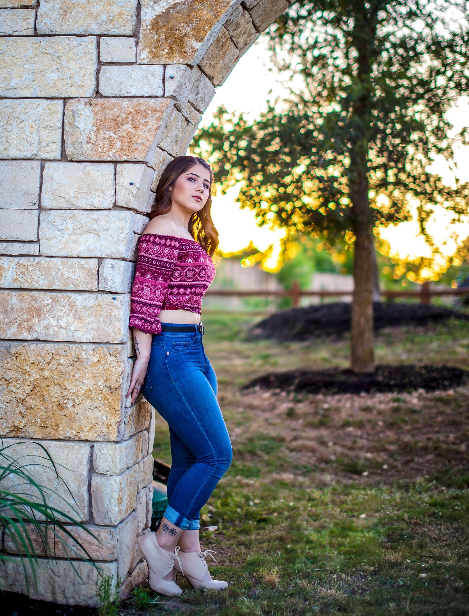 Young woman in red crop top and blue jeans leaning against stone arch outdoors at golden hour. Senior photos at sunset.