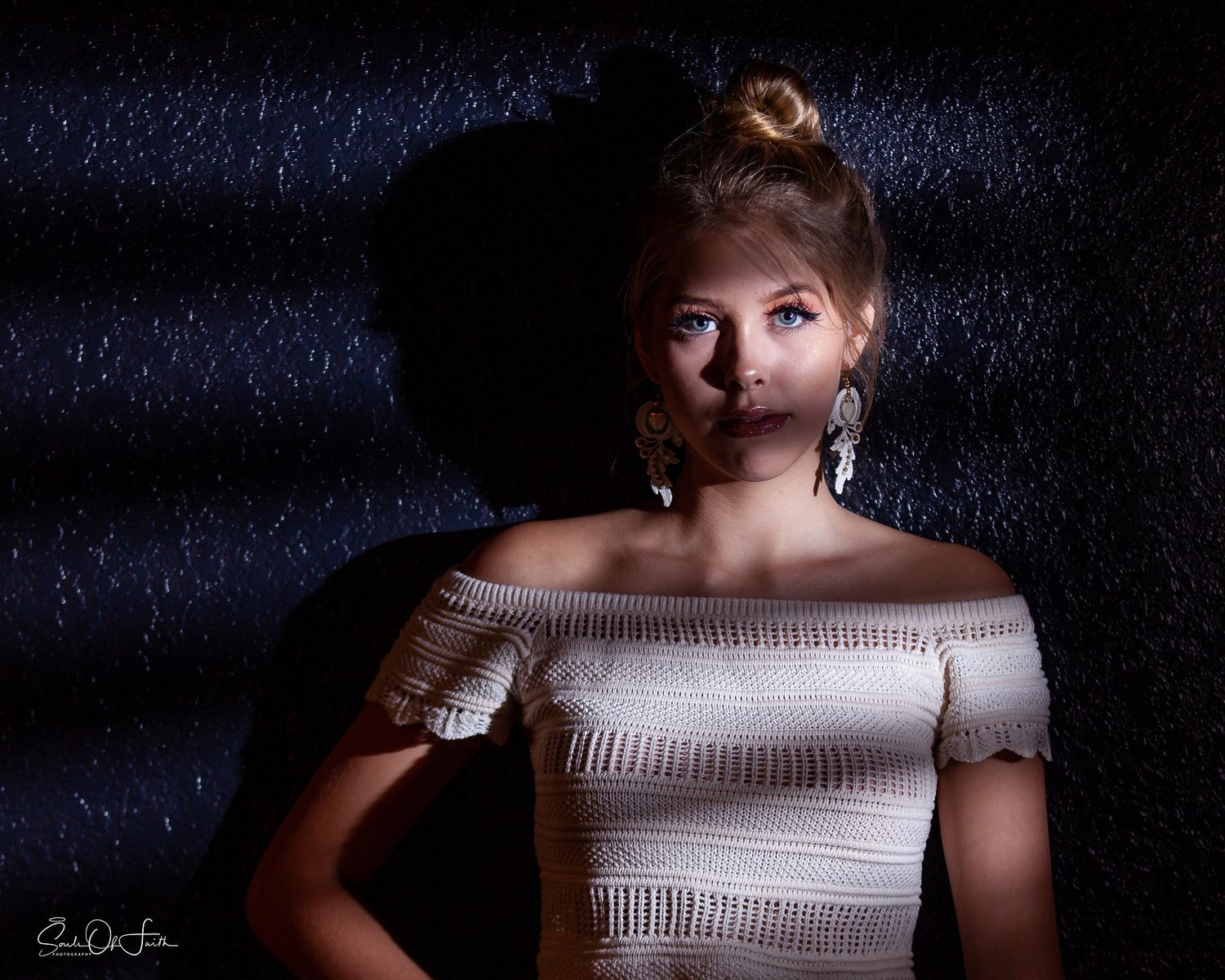 Young blonde woman in white off-shoulder top poses dramatically against dark textured backdrop wearing elegant drop earrings.