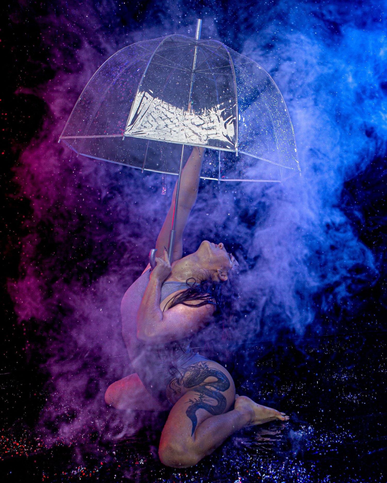 Tattooed performer holds clear umbrella amid vibrant blue and purple powder clouds in dramatic dark studio setting.