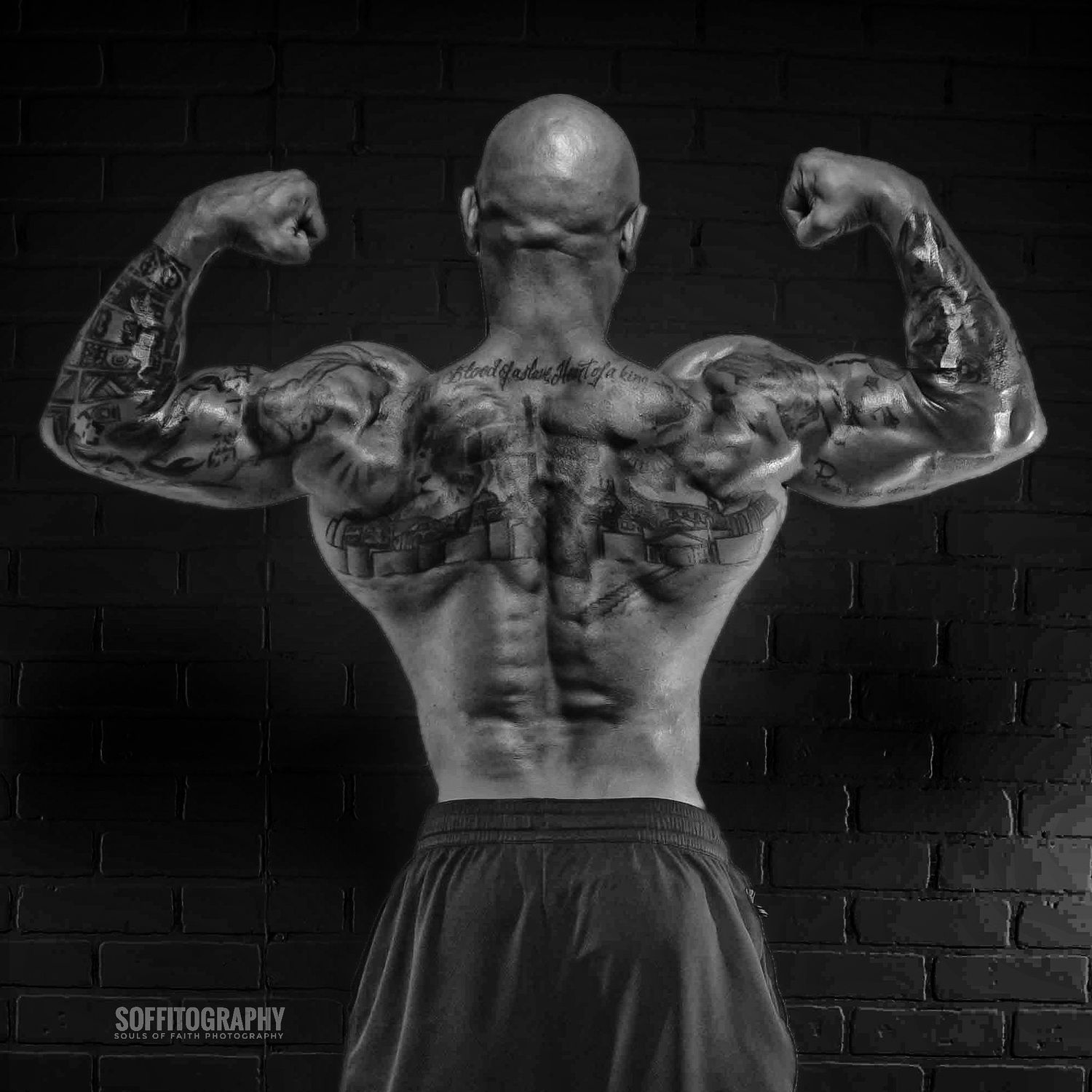 Black and white photo of a muscular tattooed man flexing his back muscles against a brick wall.