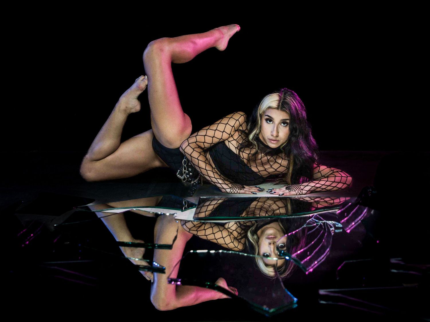 A flexible performer in fishnet bodysuit and pink tights poses dramatically on a reflective black surface.