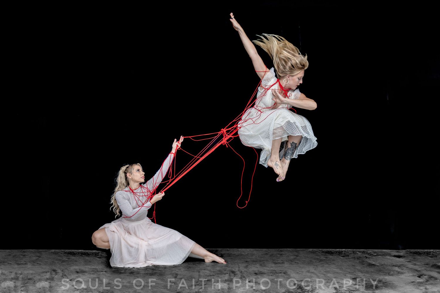Two dancers connected by red strings, one airborne and one seated, performing an artistic dance against a dark background.