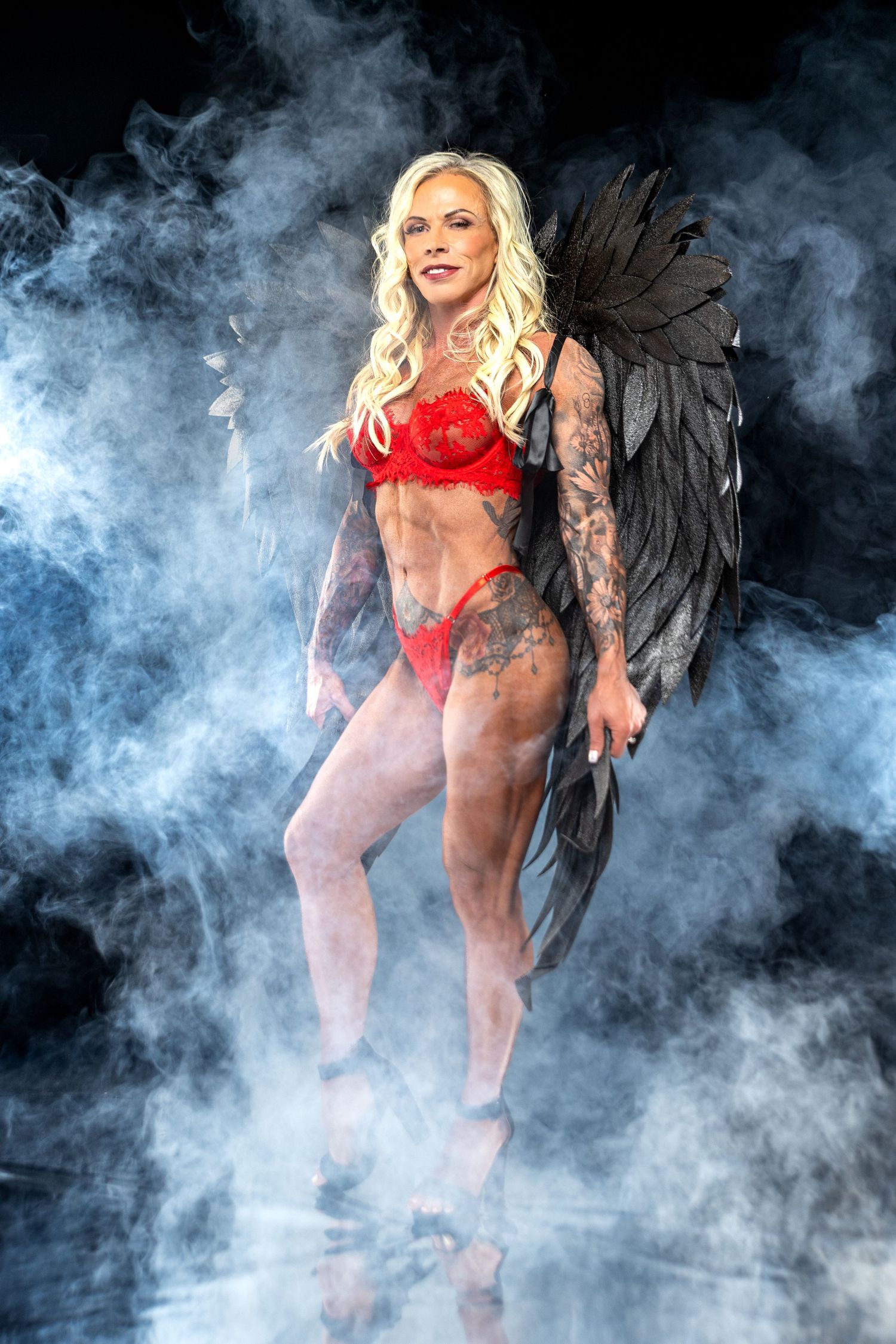 Muscular blonde woman in red lingerie with dark angel wings poses dramatically in smoky studio setting.