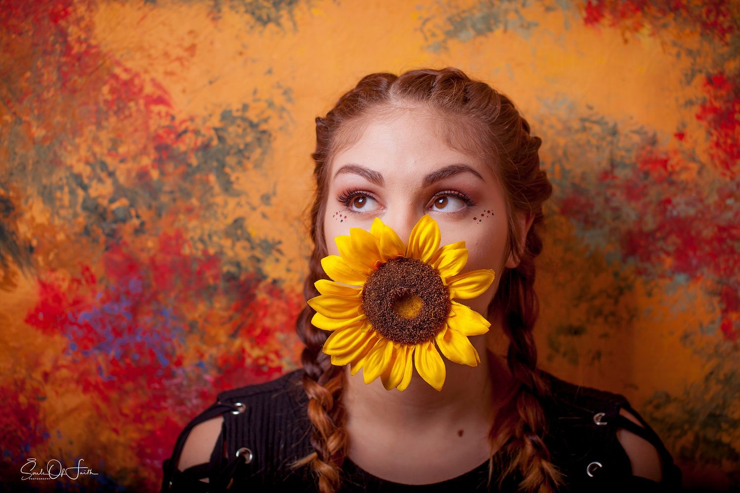 Young woman with braided hair holding a vibrant sunflower in front of her mouth against a colorful abstract background.