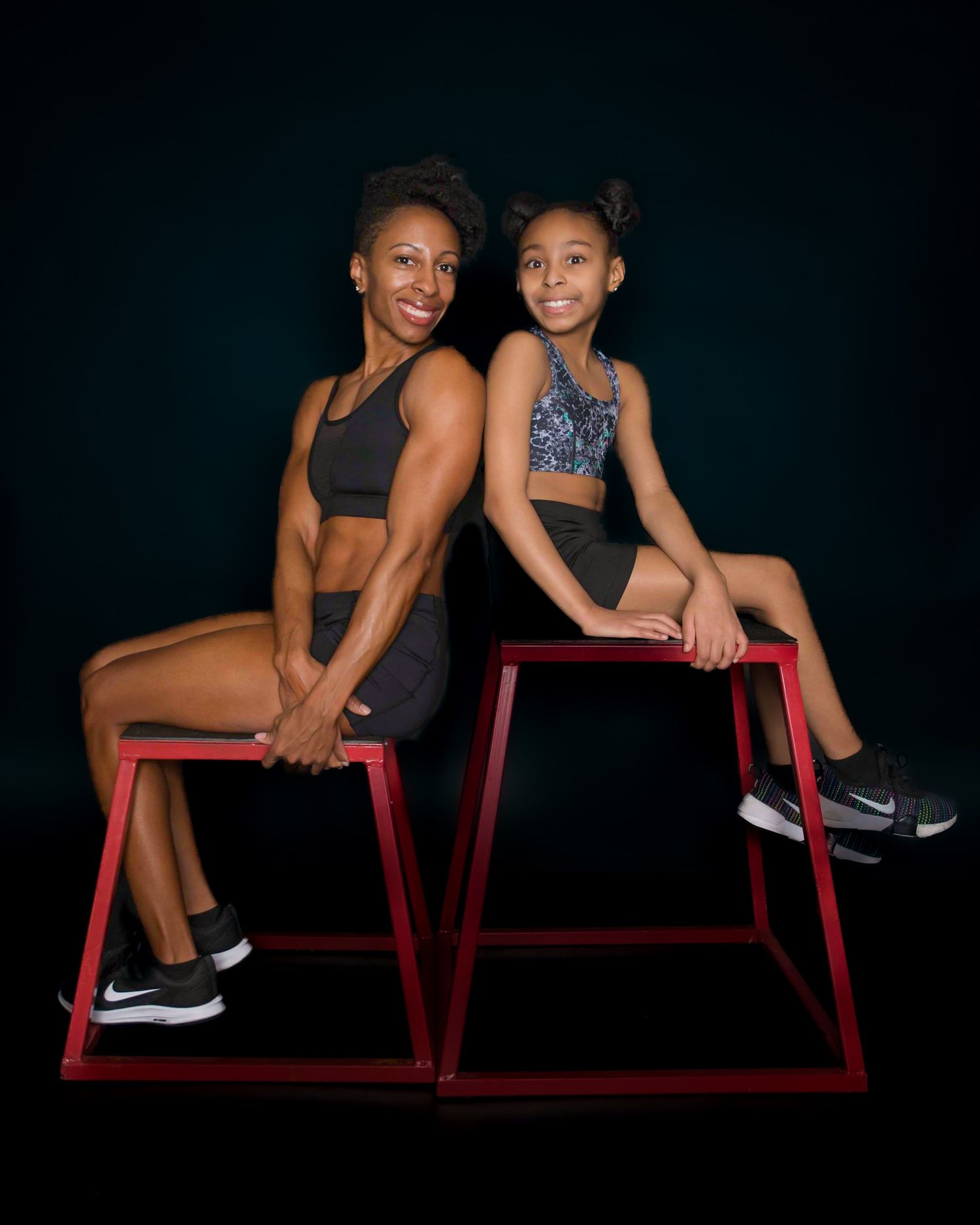 Two athletic women in sports attire pose back-to-back on red plyo boxes against a dark studio background.
