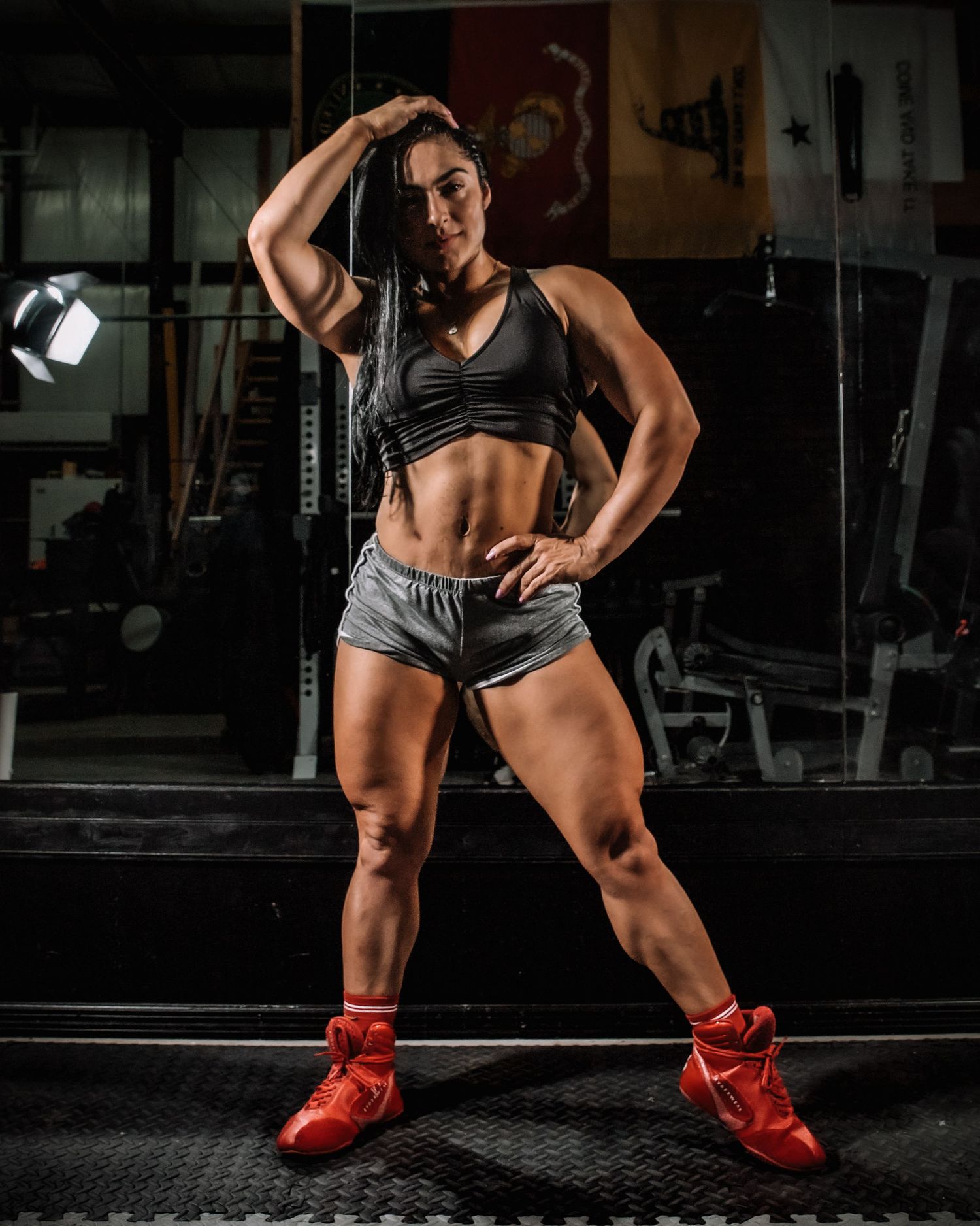 Muscular female athlete poses confidently in gym wearing sports bra, shorts, and red Nike shoes.