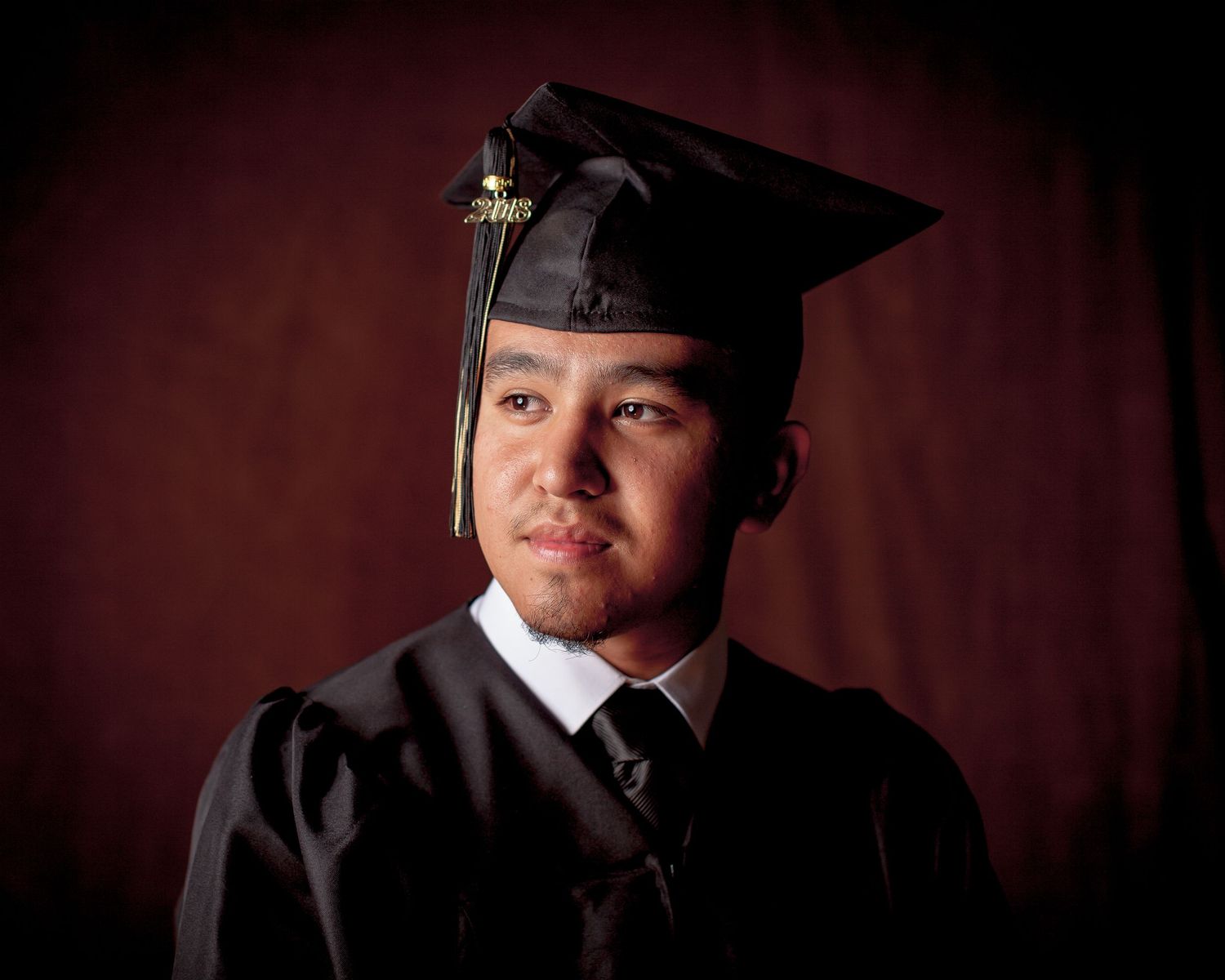 Young man in black graduation cap and gown posing for a formal portrait studio session. Graduation photos session. Studio pho