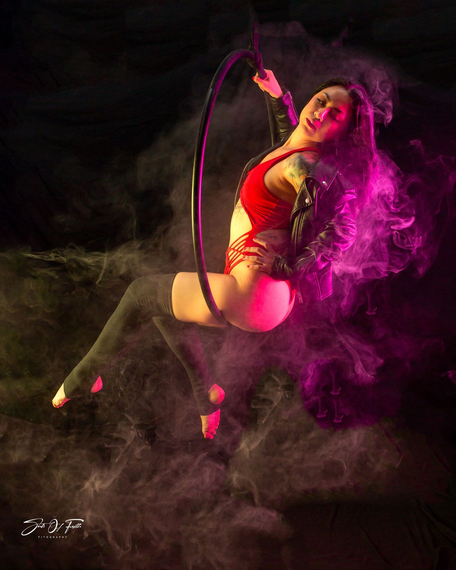 Aerial hoop performer in red leotard poses gracefully surrounded by vibrant pink and purple smoke on dark background.