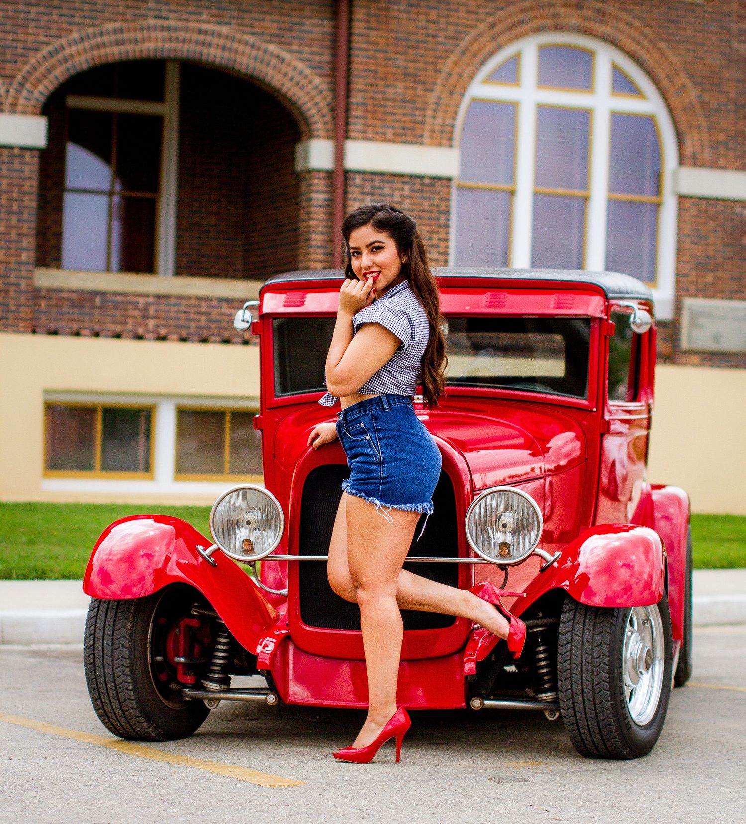 Young woman in denim shorts and red heels posing with a classic red hot rod car in front of a brick building. pinup coca cola