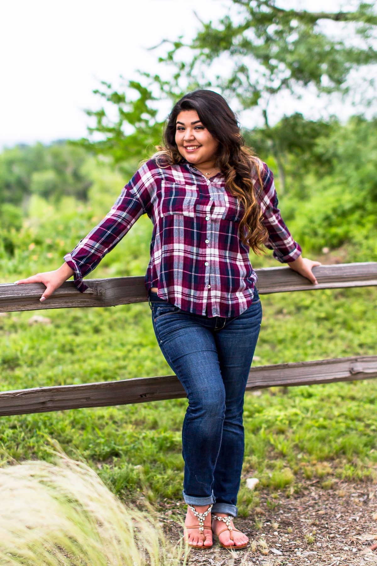 Young woman in plaid shirt and jeans leaning on wooden fence outdoors with green trees in background.
