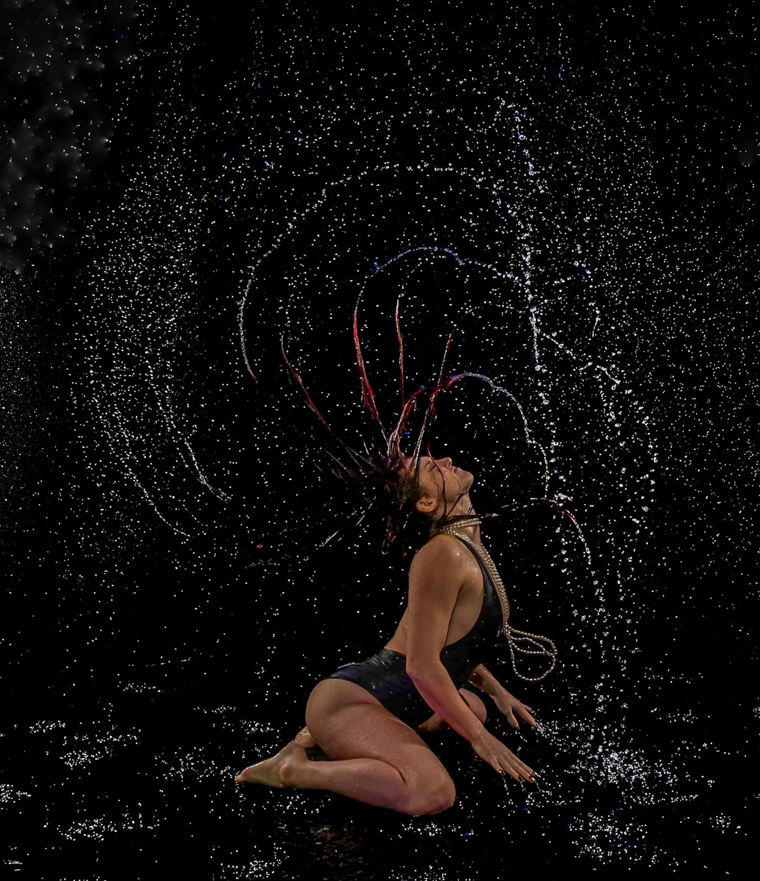 Performer kneeling in water, flipping colorful braids creating arc of droplets against dark background.