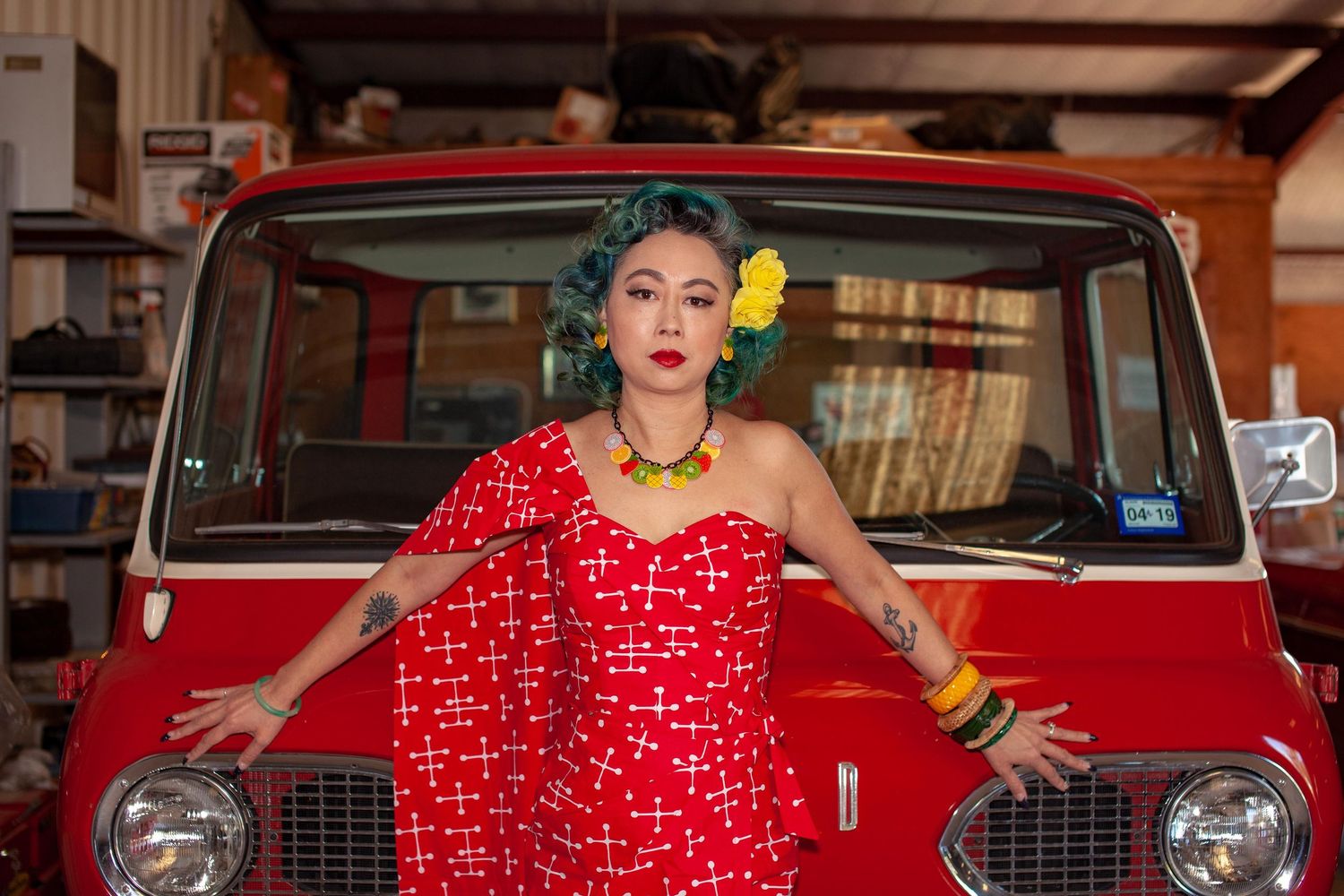 Woman in red polka dot dress with teal hair and yellow flower poses in front of a vintage red van in a garage.