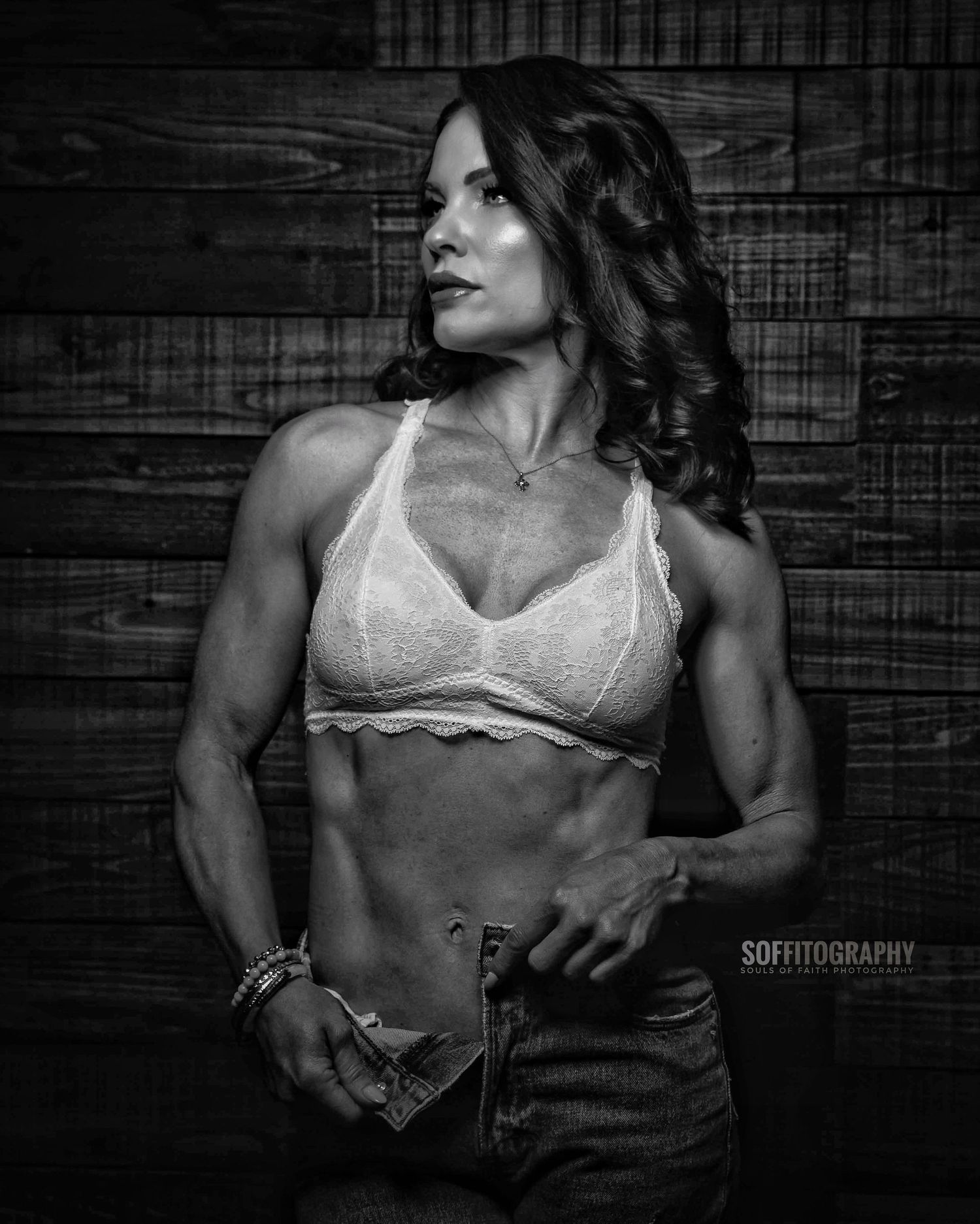Black and white photo of a fit woman in a lace bralette and jeans posing against a wooden wall backdrop.