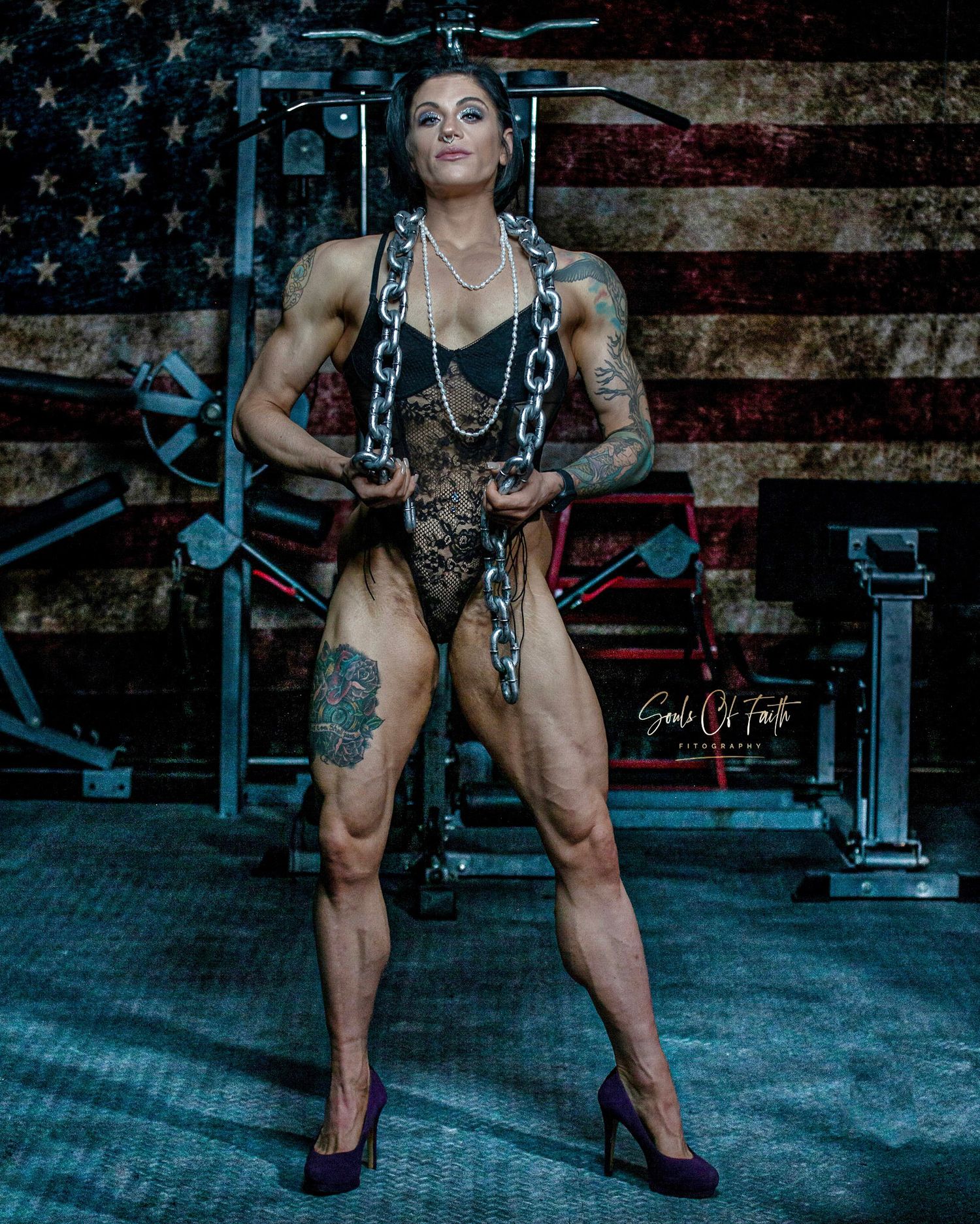 Muscular woman in lace outfit and heels poses with chains in a gym before an American flag backdrop.
