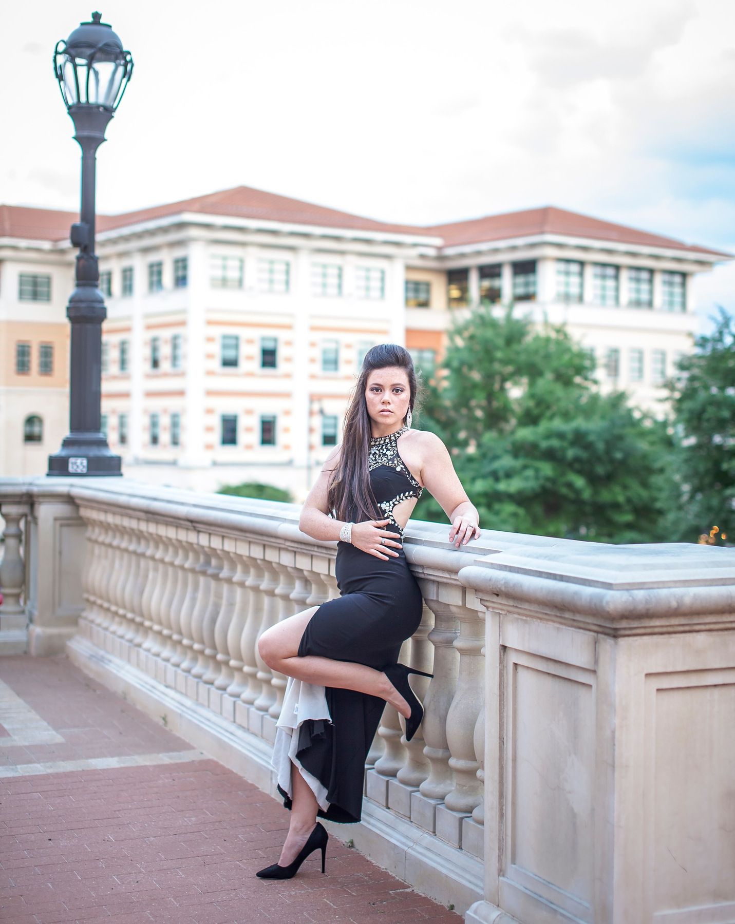 Woman in black halter dress and heels poses on stone balcony railing with elegant building backdrop.