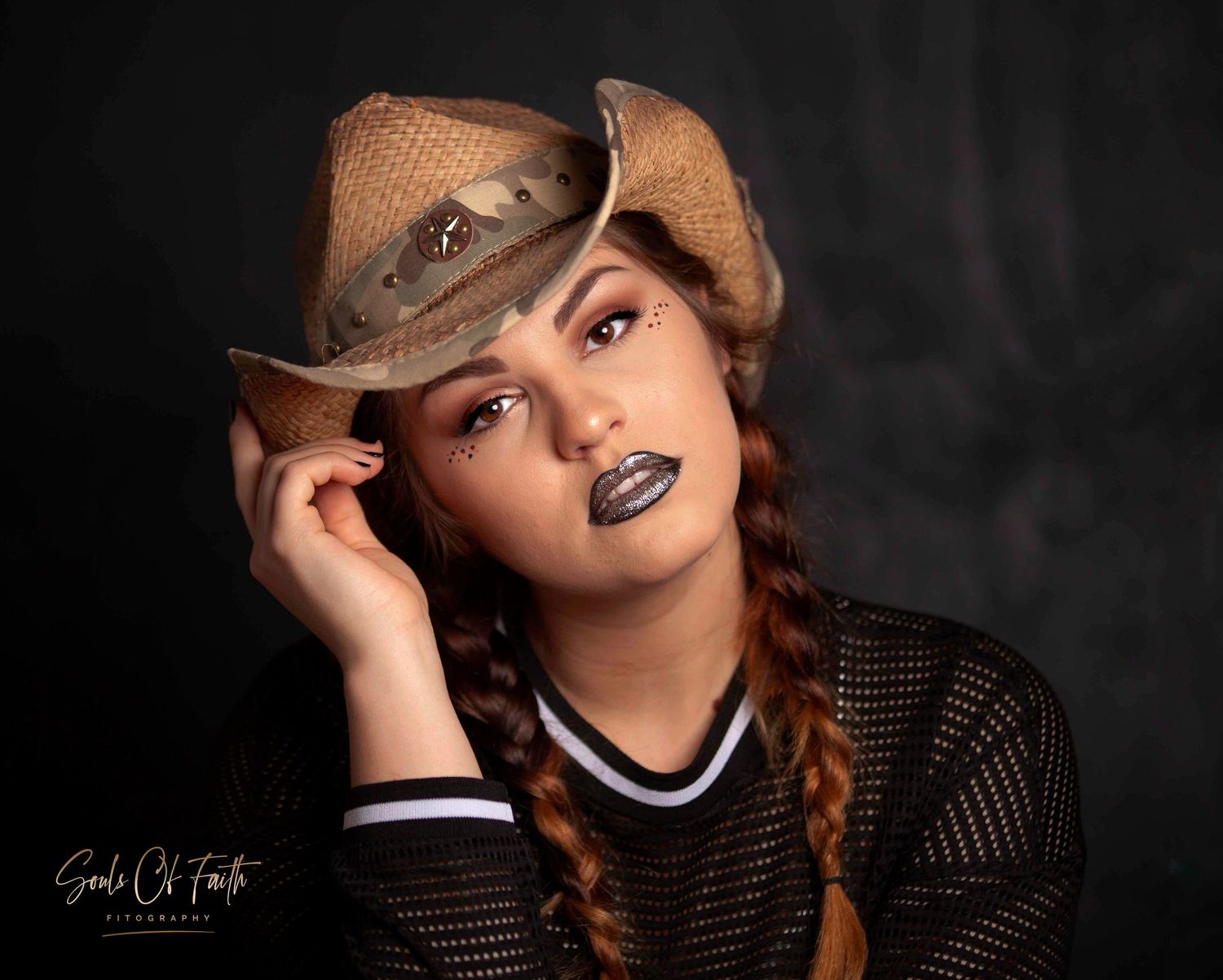 Young woman in a burlap cowboy hat and mesh top with dark lipstick and braids poses for a studio portrait.