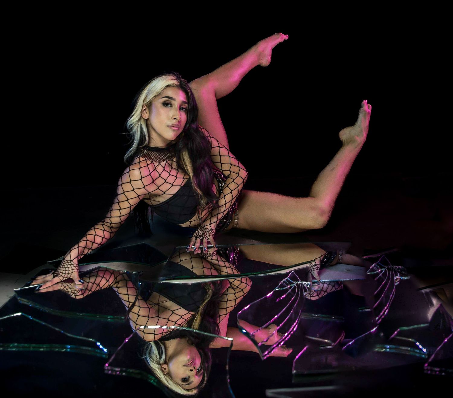 Dancer in fishnet bodysuit poses dramatically on reflective surface with leg raised, lit by pink and green stage lighting.