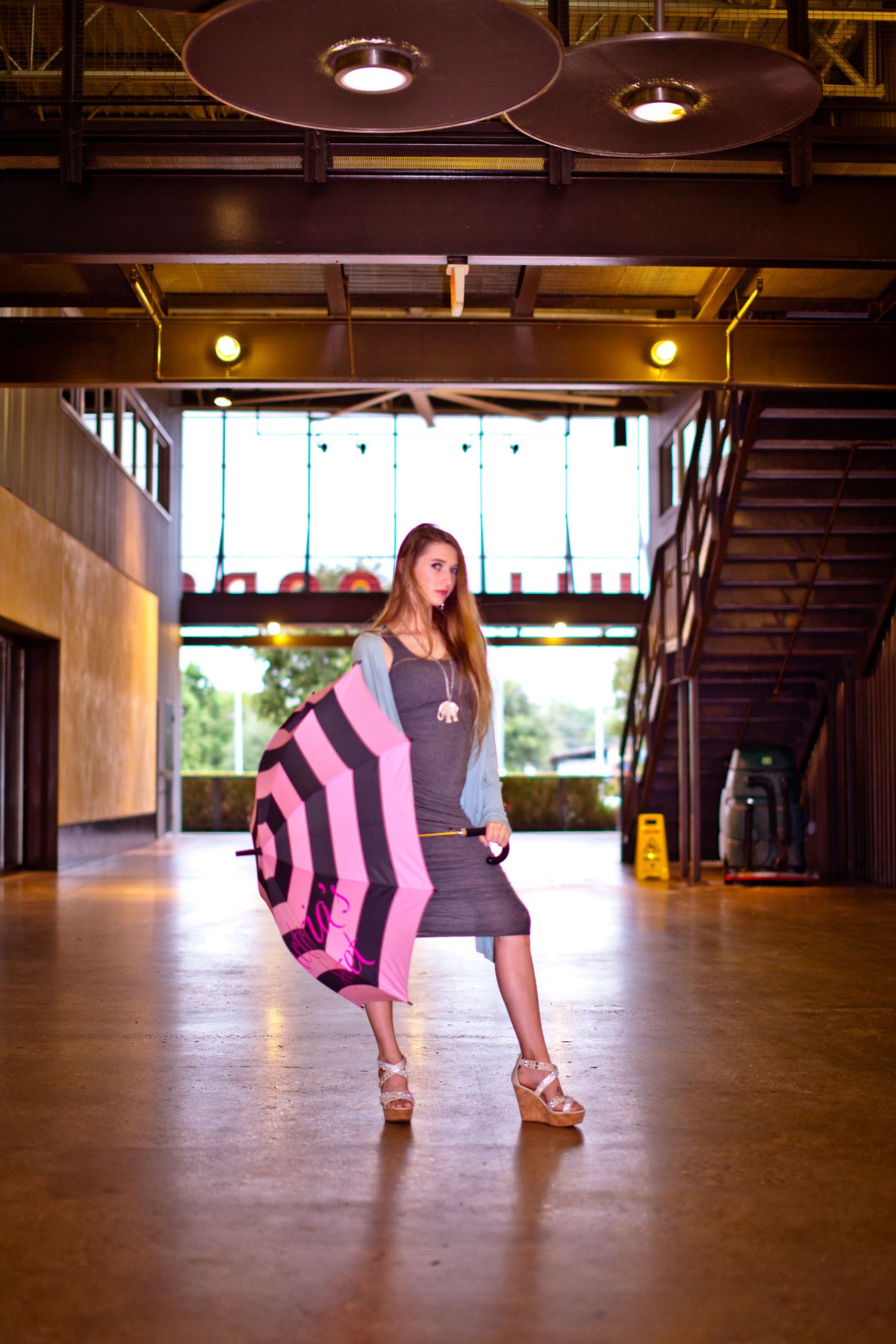 Young woman in gray dress holding pink striped umbrella poses in modern industrial building interior.