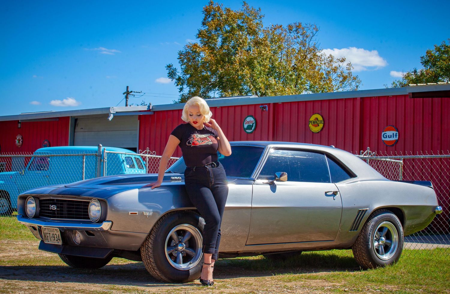Blonde woman leaning on a classic silver Camaro muscle car at an outdoor car show on a sunny day.SATX ATX HTX DFW Austin San Antonio Houston