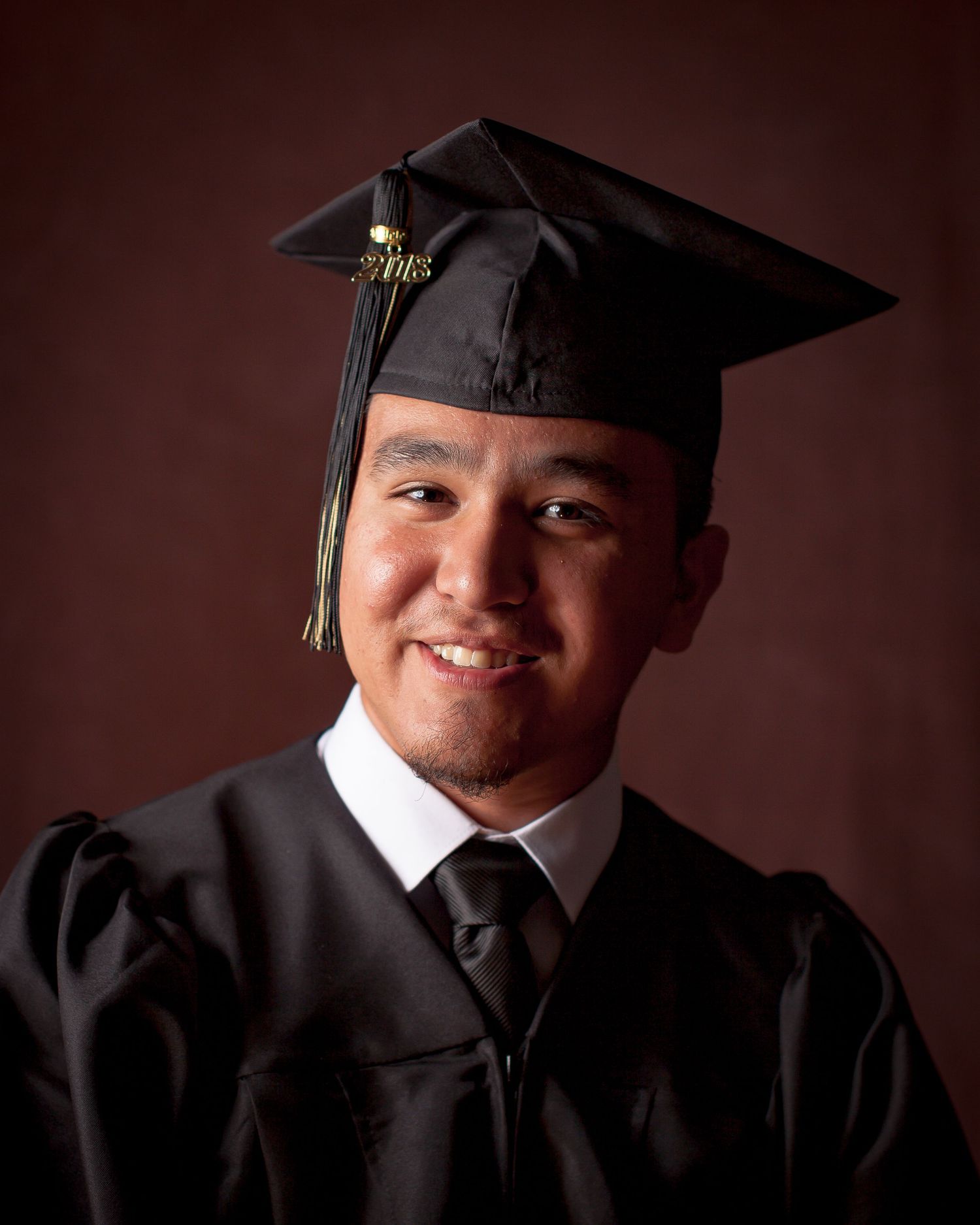 Young man smiling in black graduation cap and gown, posing for a formal graduation portrait photo.