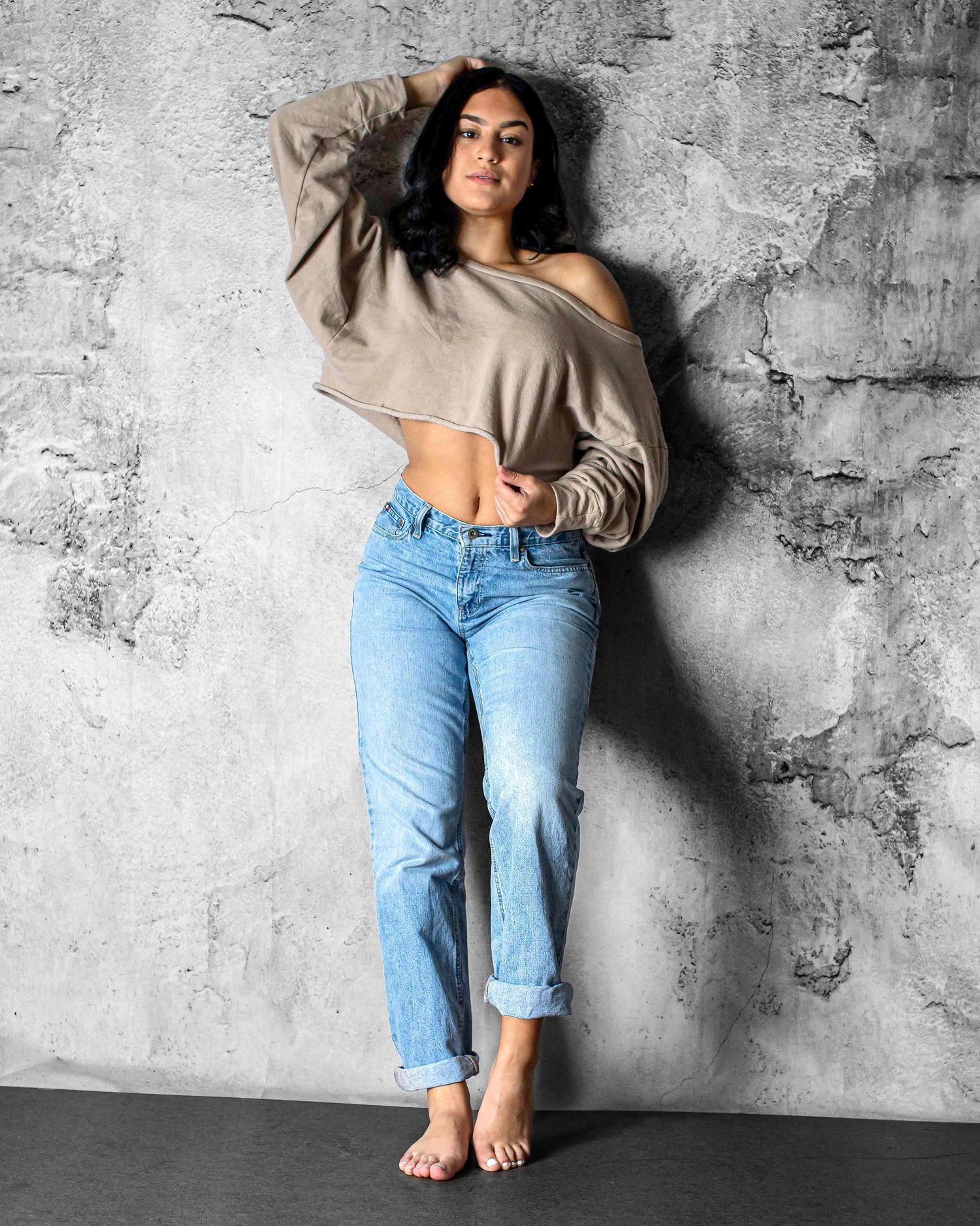 Woman in beige off-shoulder top and light blue jeans posing barefoot against a gray concrete wall.