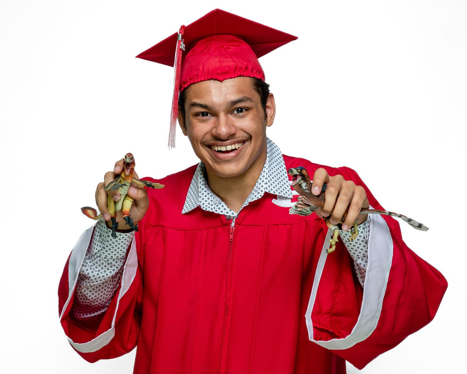 Smiling young man in red graduation cap and gown holding diplomas, celebrating academic achievement against white background.