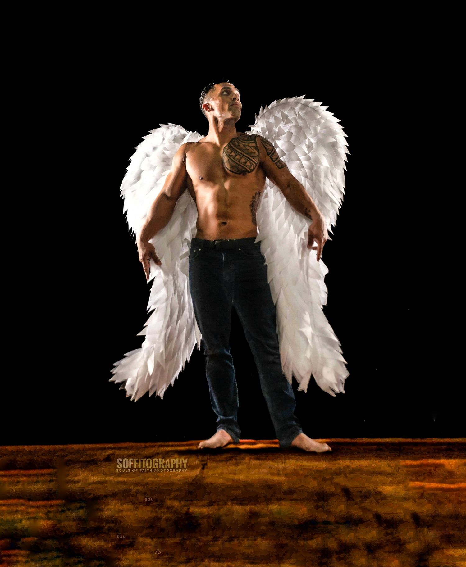 Shirtless man with large white angel wings poses dramatically against a dark background wearing jeans.