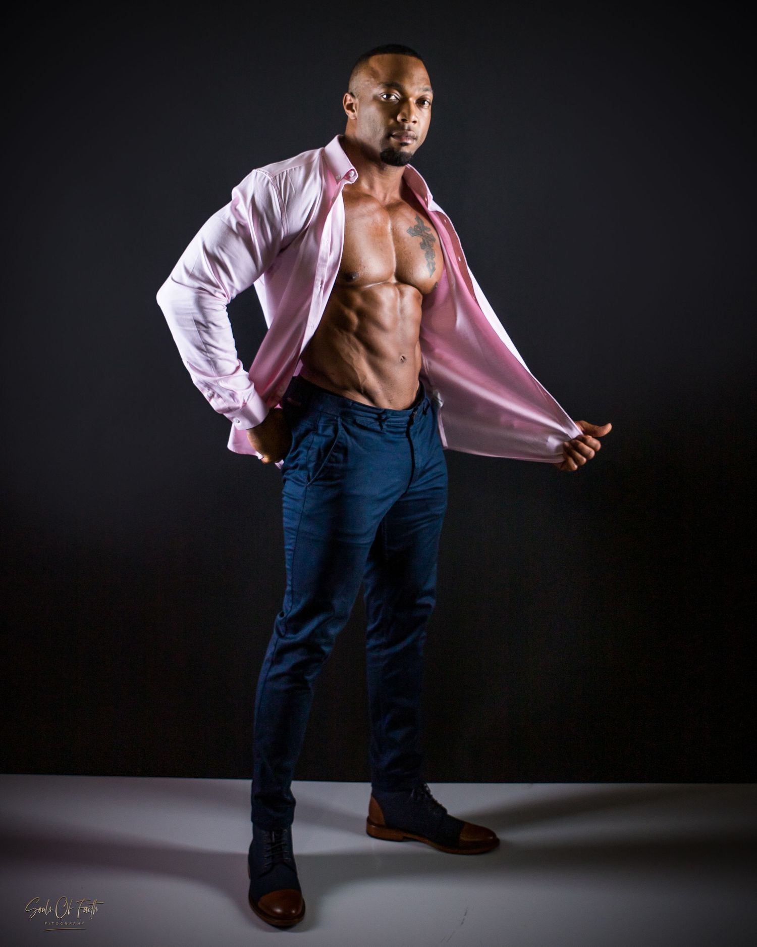 A muscular man poses confidently in an open pink shirt and blue jeans, showcasing his toned physique against a dark studio background.