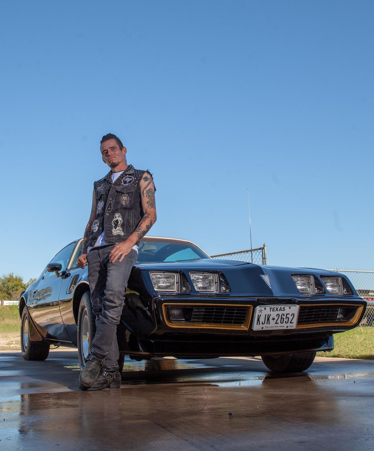 Man in vest leaning on black Pontiac Firebird with Texas license plate KJX-2652 under clear blue sky.