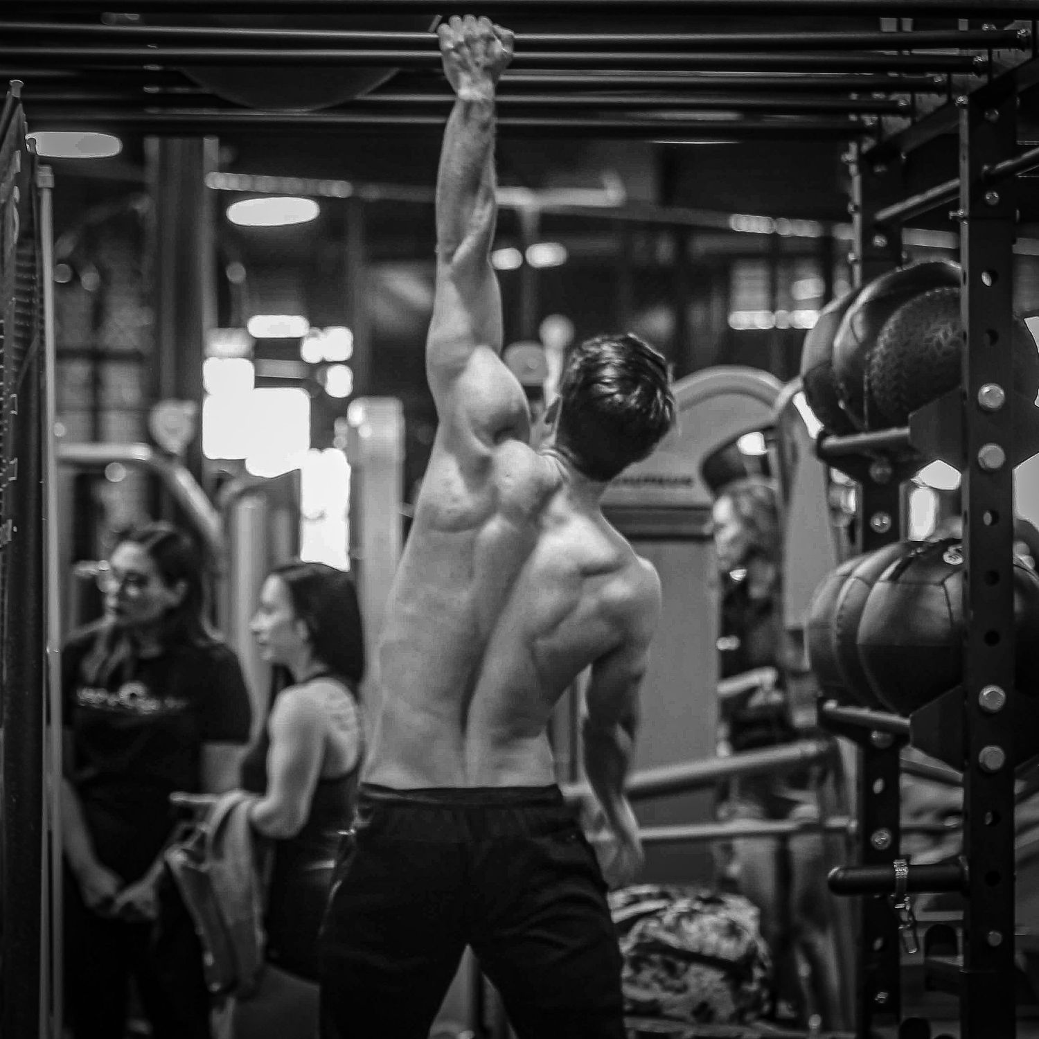 Shirtless man doing pull-ups on a bar in a busy gym, black and white fitness photography.