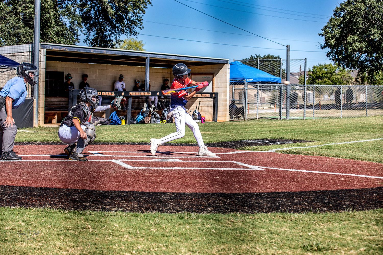 Baseball batter swings at home plate during a sunny outdoor game, with catcher and umpire behind him.