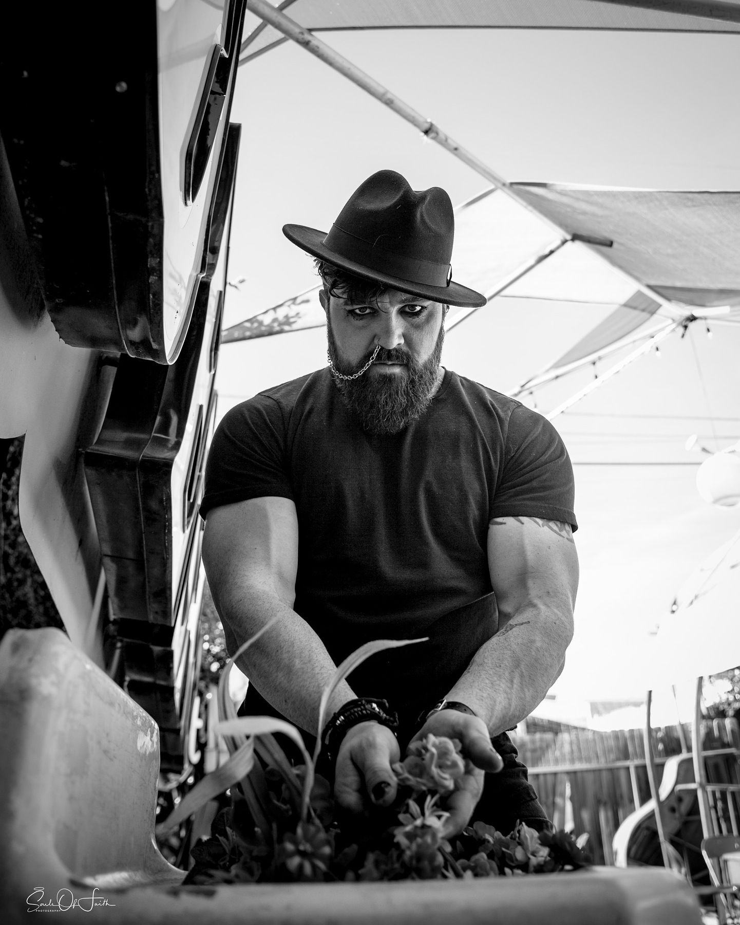 Black and white photo of a bearded man in a wide-brim hat and black t-shirt working with flowers outdoors.