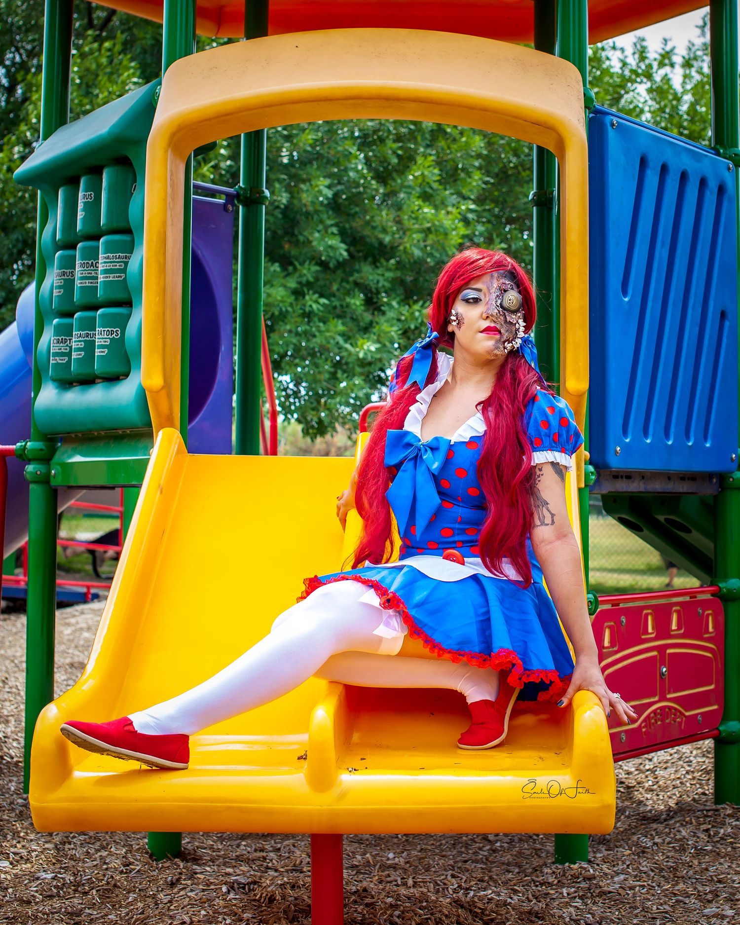 Woman in Raggedy Ann costume with red wig and face paint sits on yellow playground slide at colorful play structure. cosplay makeup