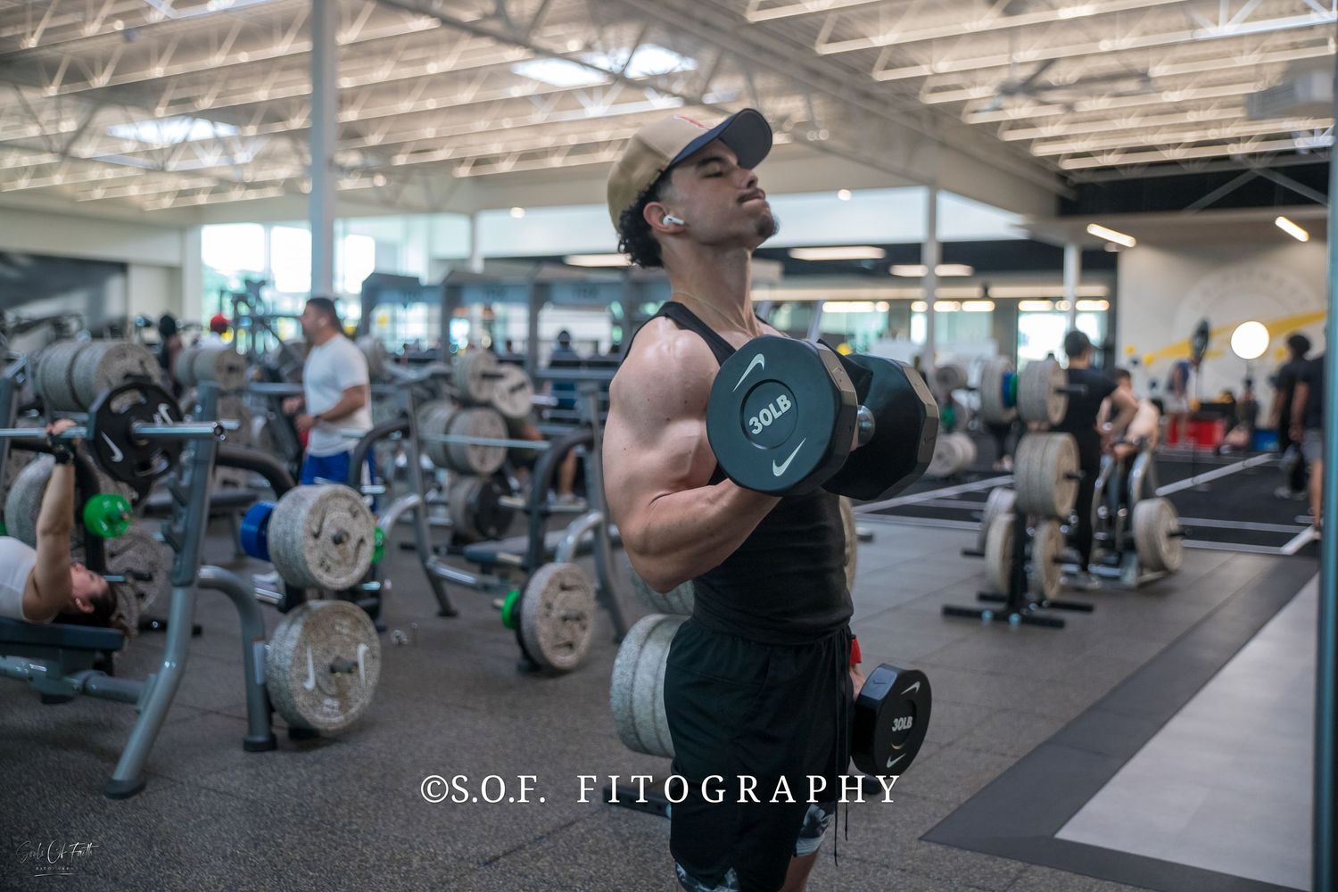 Muscular man in a hat doing dumbbell curls in a modern gym, showcasing strength training and fitness.
