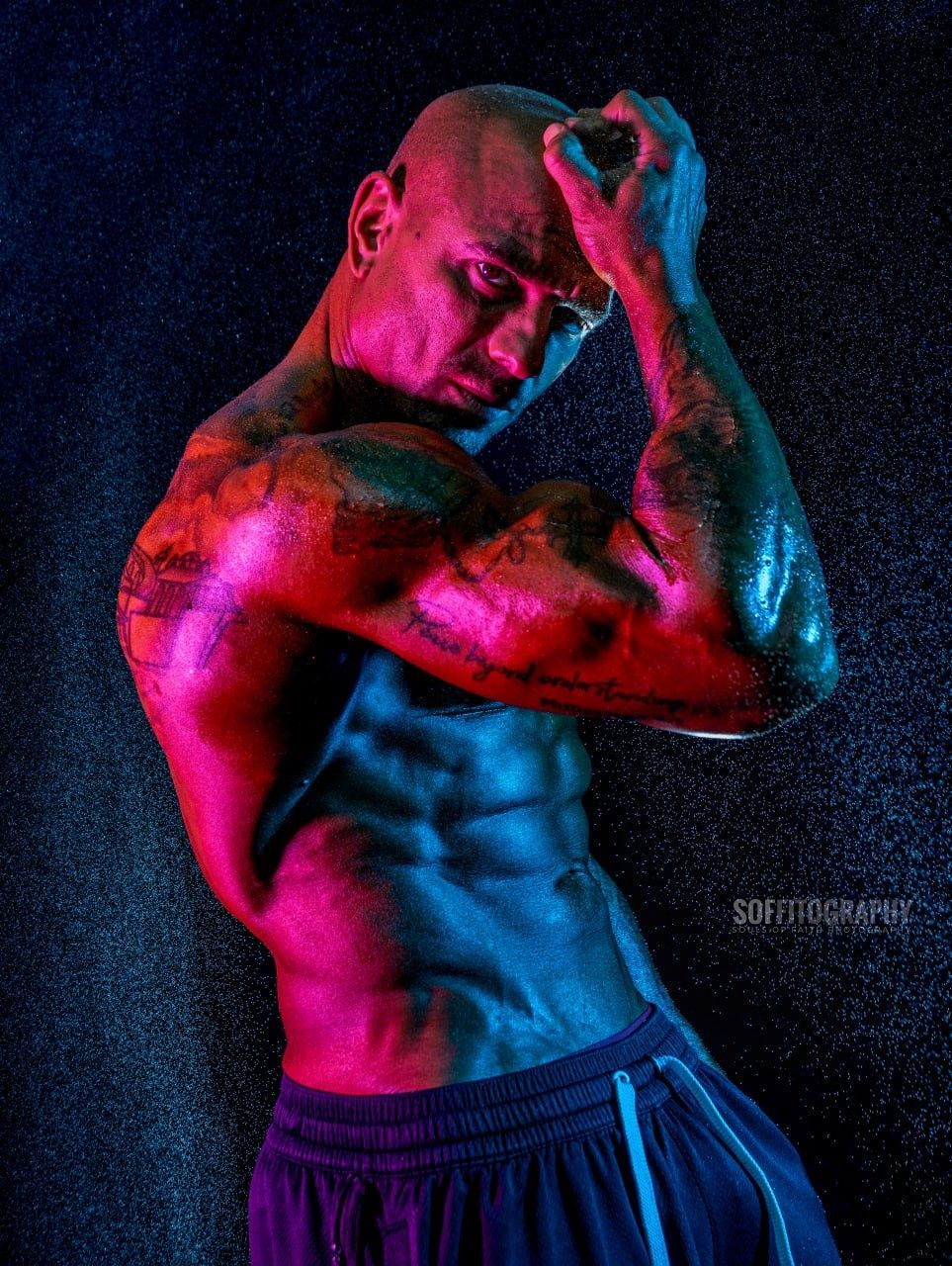 Muscular bald man with tattoos poses shirtless under dramatic red and blue lighting in a professional fitness photo.