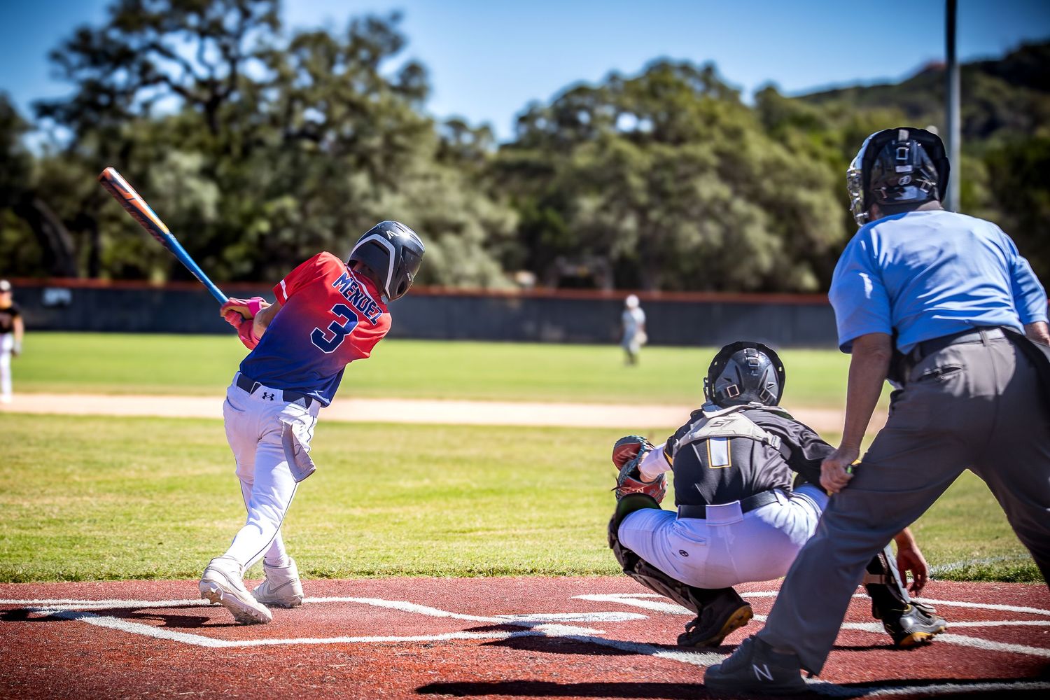 Baseball batter in red uniform swings at pitch with catcher and umpire behind home plate on sunny day.