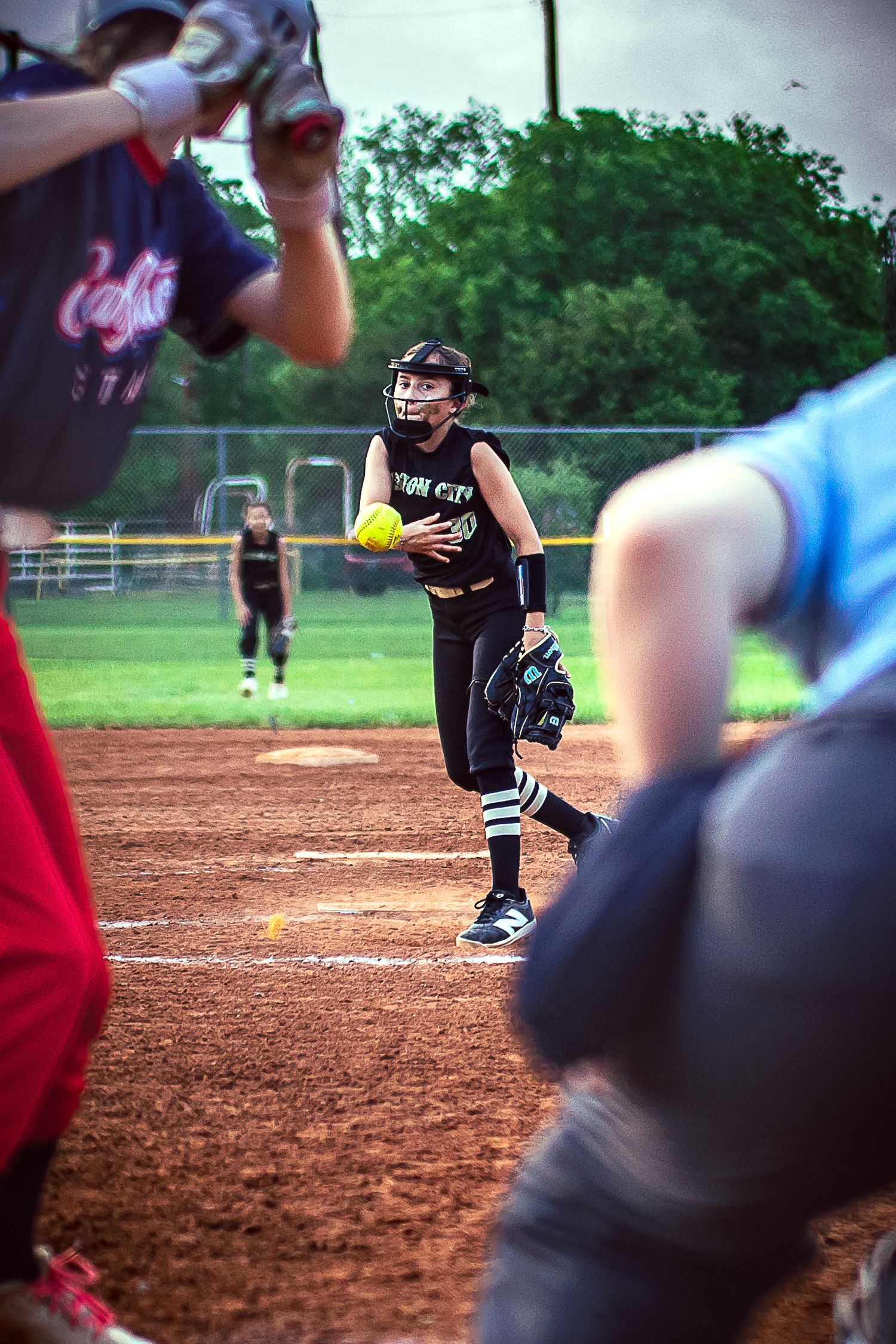 Young softball pitcher in black uniform winds up to throw a yellow ball during a youth league game on a dirt infield.
