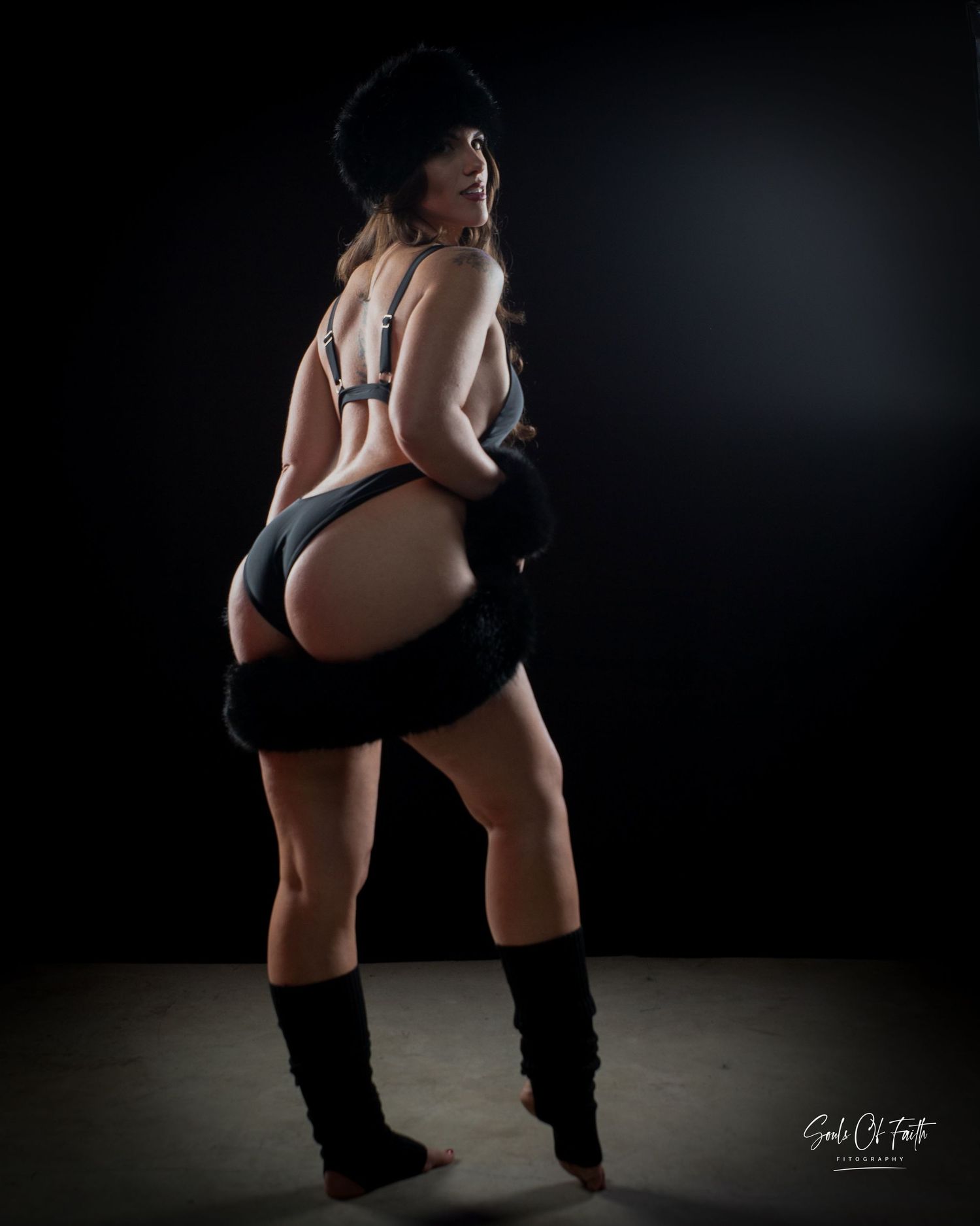 Woman posing in dark studio wearing black bodysuit, leg warmers, and hat in dramatic low-key lighting.