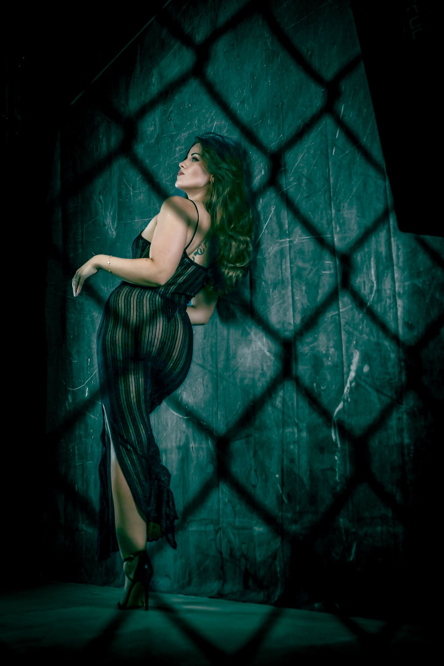 A woman in a sheer striped dress poses against a dark teal wall, viewed through a chain-link fence.